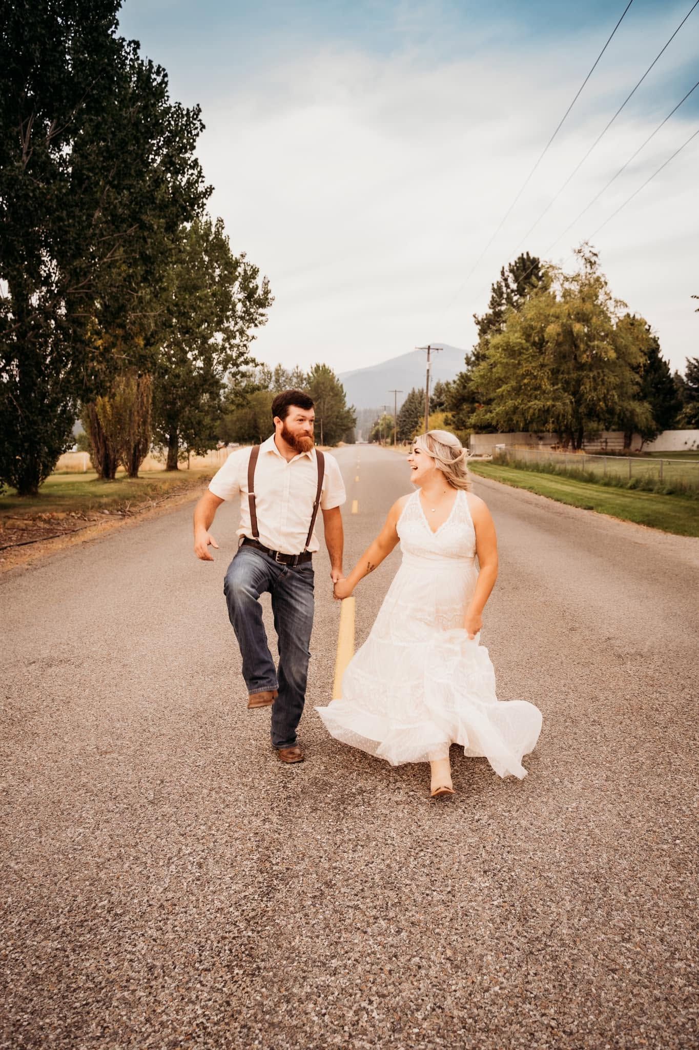 Bride and groom holding hands walking down country road, bride in lace dress, groom in suspenders.