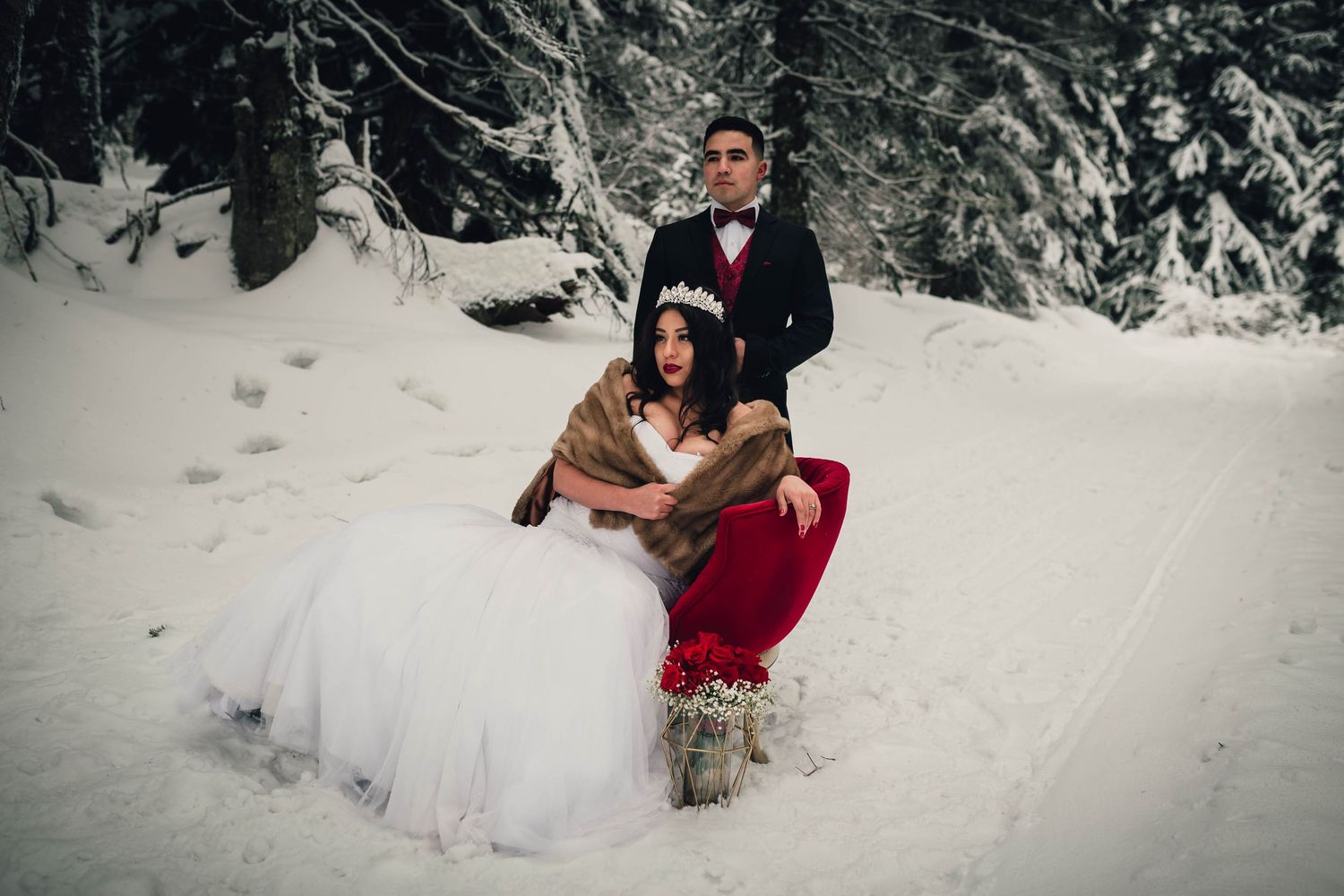 Bride in white gown with fur wrap sits on red chair in snowy winter forest with groom in black tuxedo.