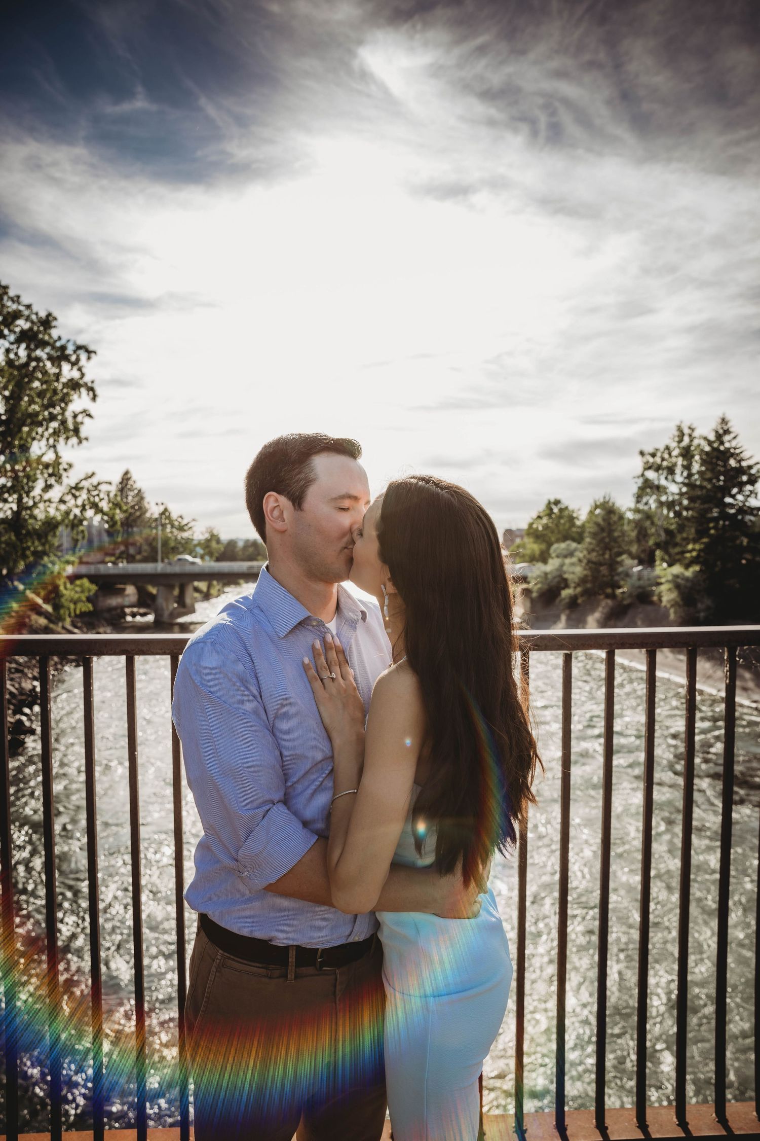 Couple kissing on bridge over river in golden sunlight with rainbow lens flare.