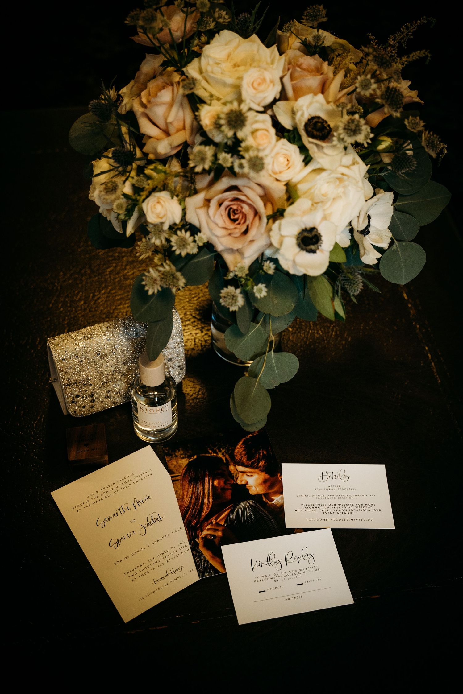 Wedding flat lay featuring floral bouquet with roses, anemones, eucalyptus, invitation suite and perfume on dark surface.