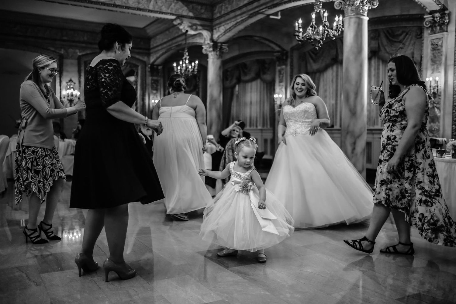 Flower girl twirls on dance floor surrounded by wedding guests in grand ballroom.