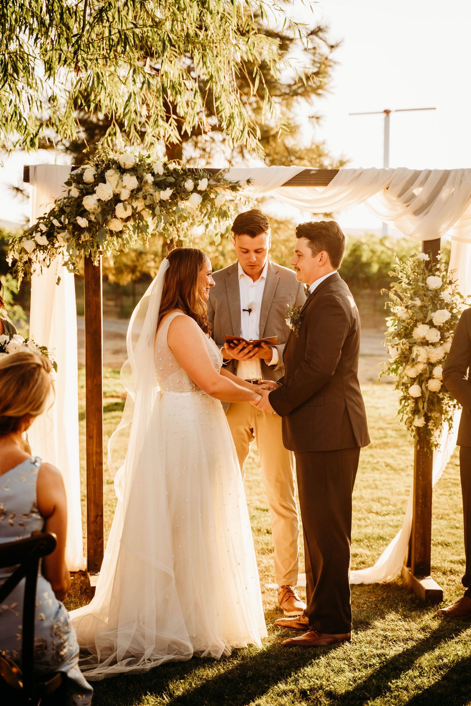 Bride and groom exchange vows under a floral arch with white roses and greenery at an outdoor wedding ceremony.