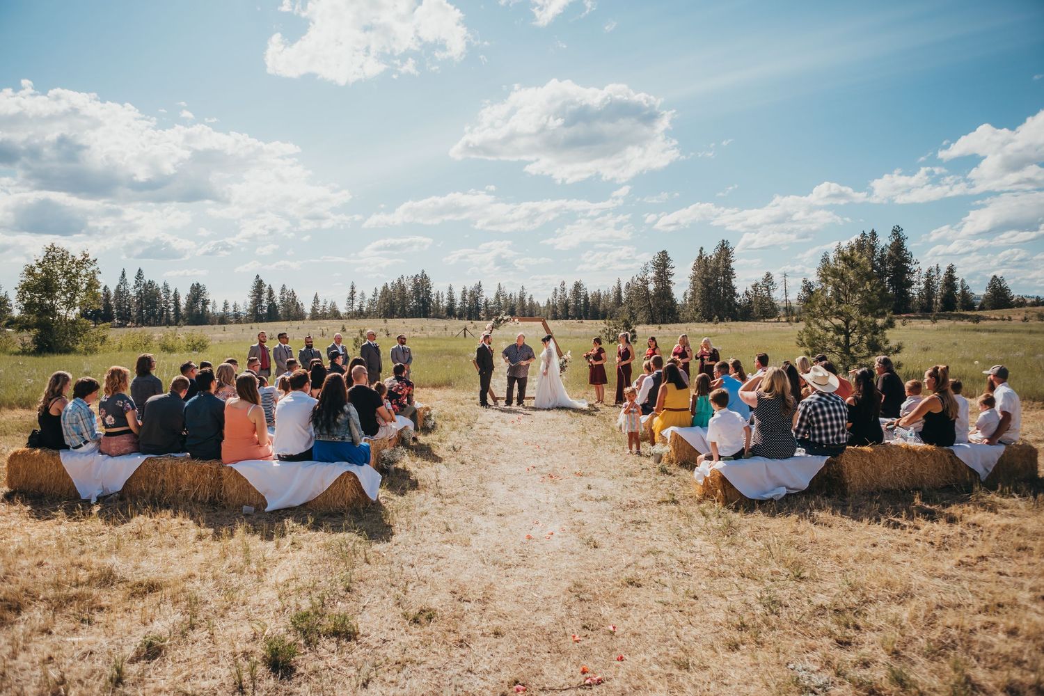 Outdoor rustic wedding ceremony with guests seated on hay bales in a golden field surrounded by trees.