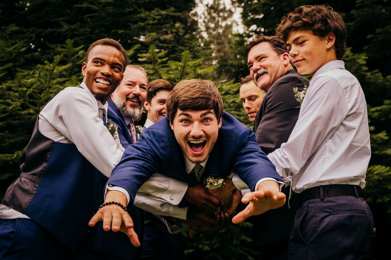 Excited groom in a blue suit surrounded by laughing groomsmen in a playful outdoor wedding photo.