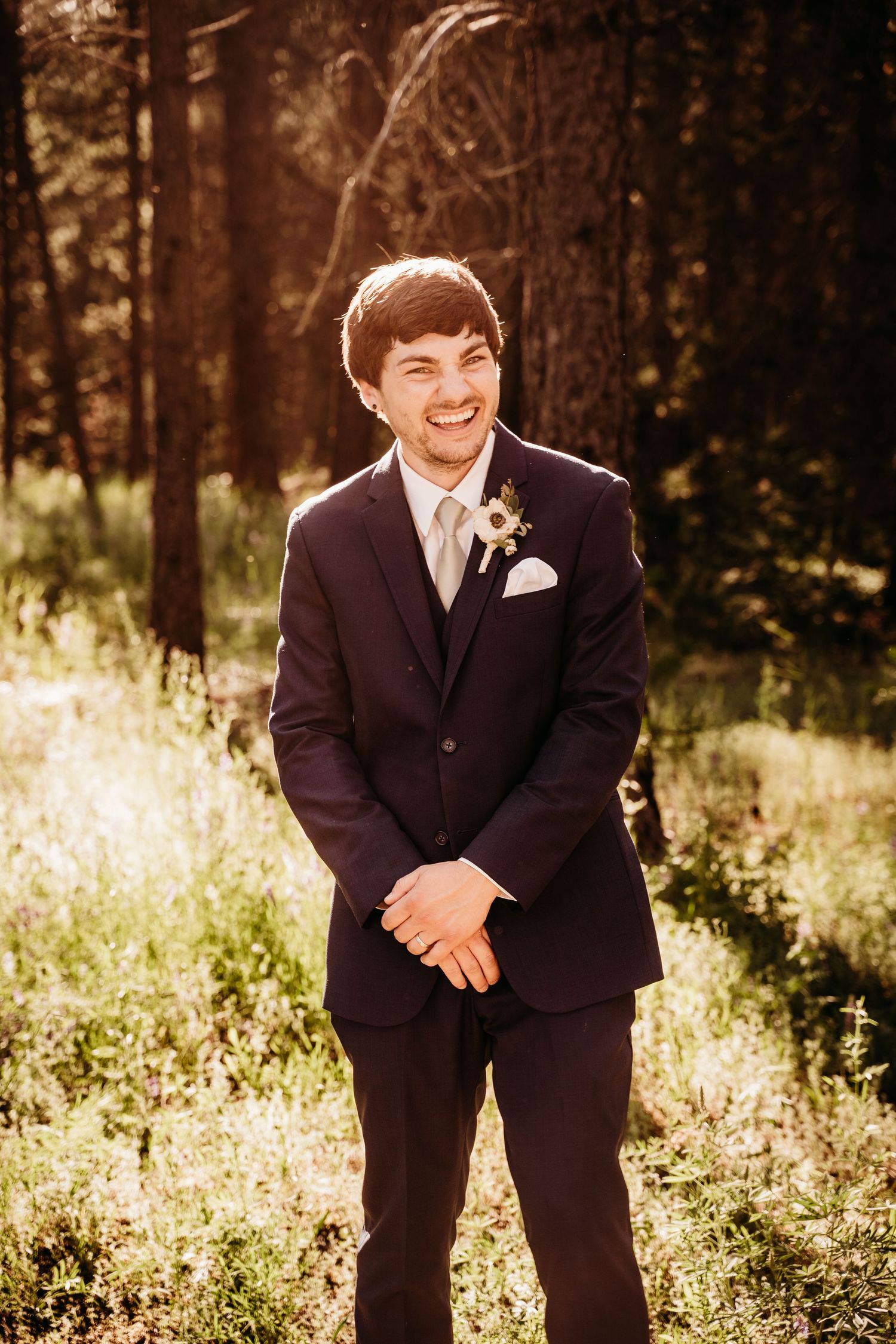 Smiling groom in navy suit with boutonniere stands in sunlit forest clearing on his wedding day.