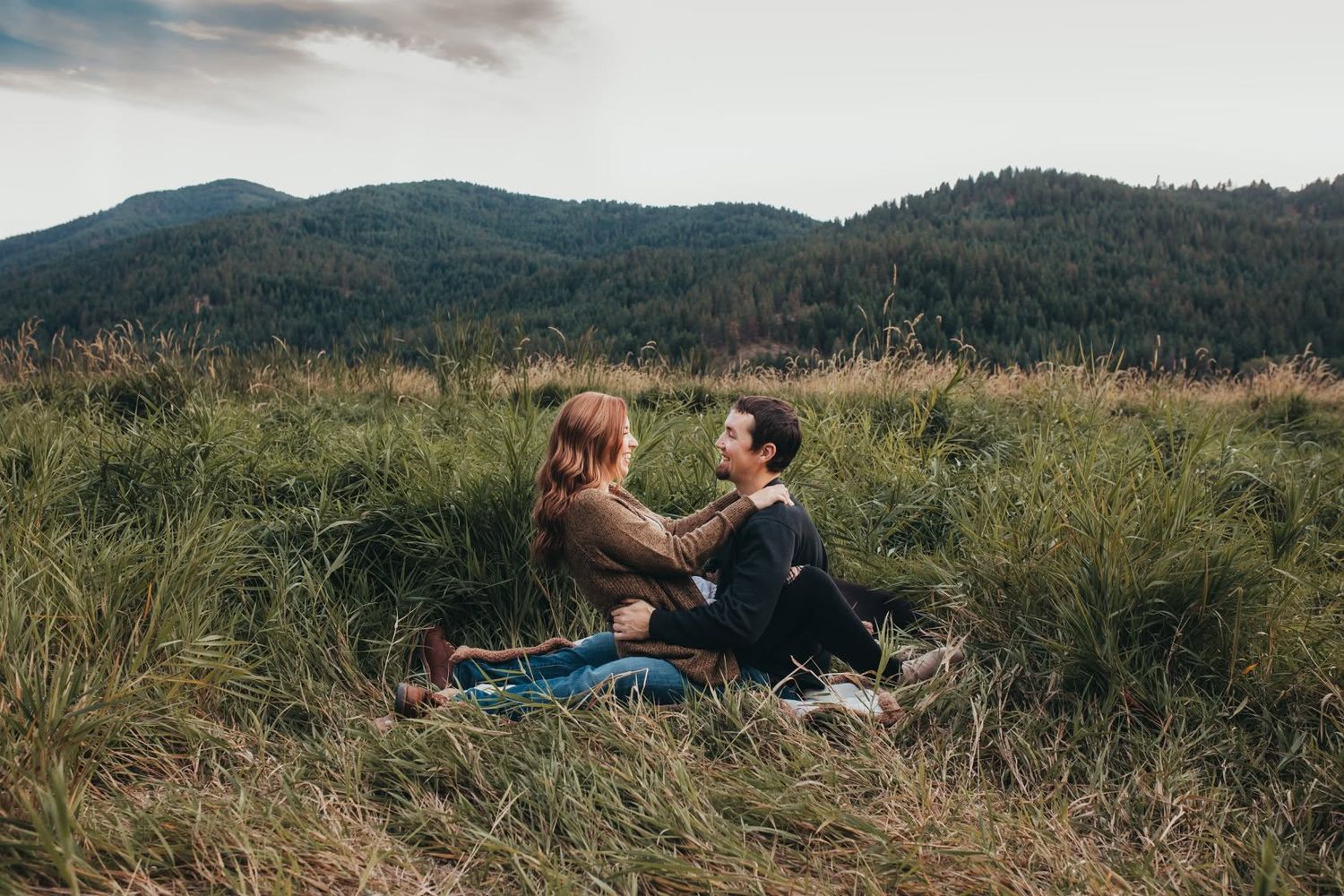 A couple sits together in a grassy meadow facing each other with rolling mountains visible in the background.