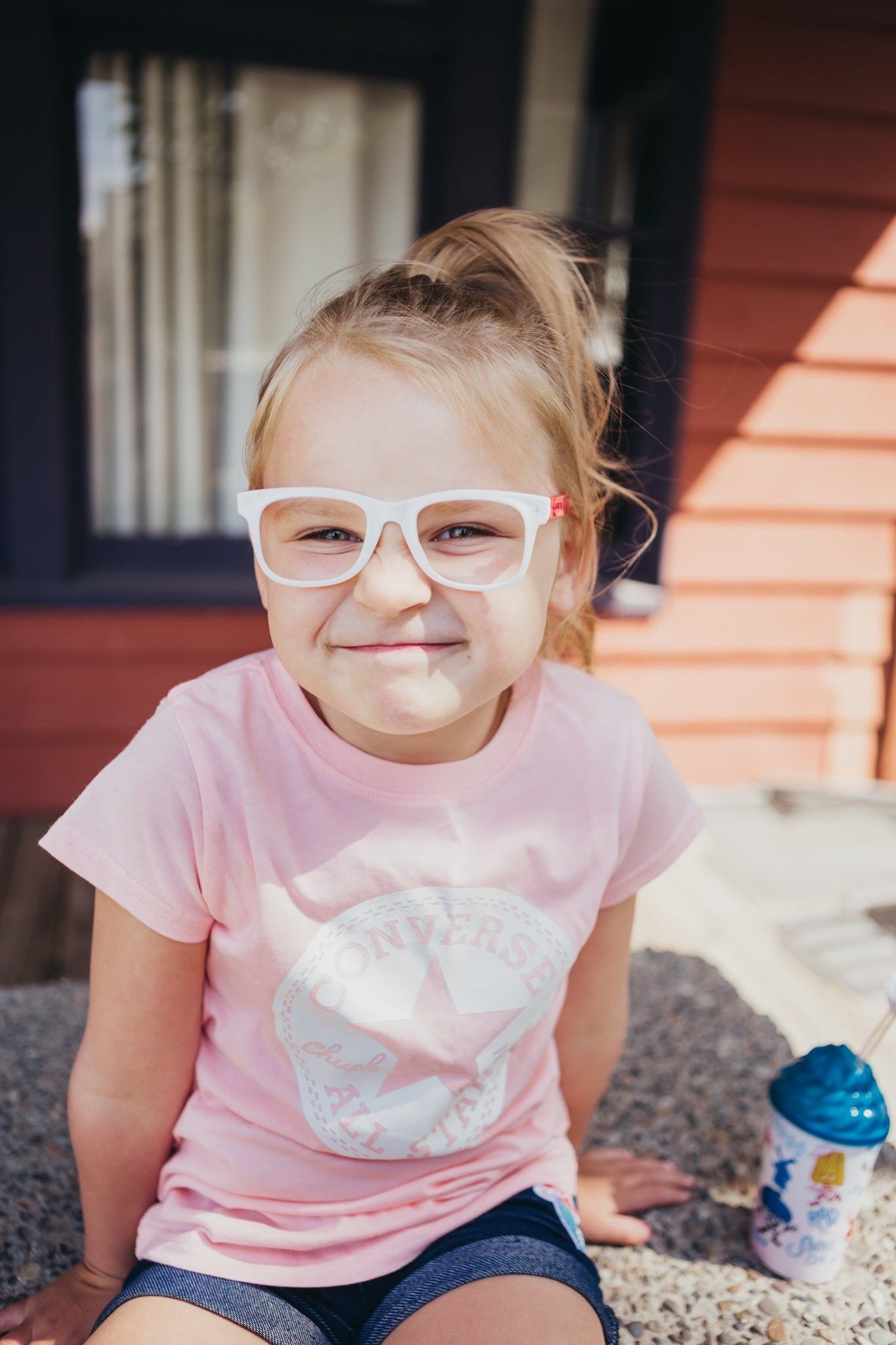 Smiling young girl wearing white-framed glasses and a pink Converse Chuck Taylor t-shirt sitting outdoors.