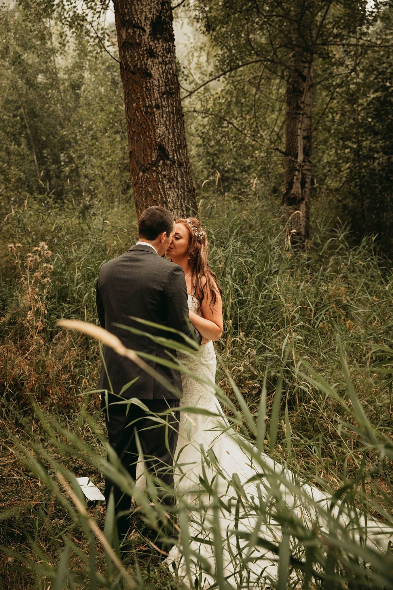 Bride and groom share a romantic kiss surrounded by tall green grass and trees in a forest setting.