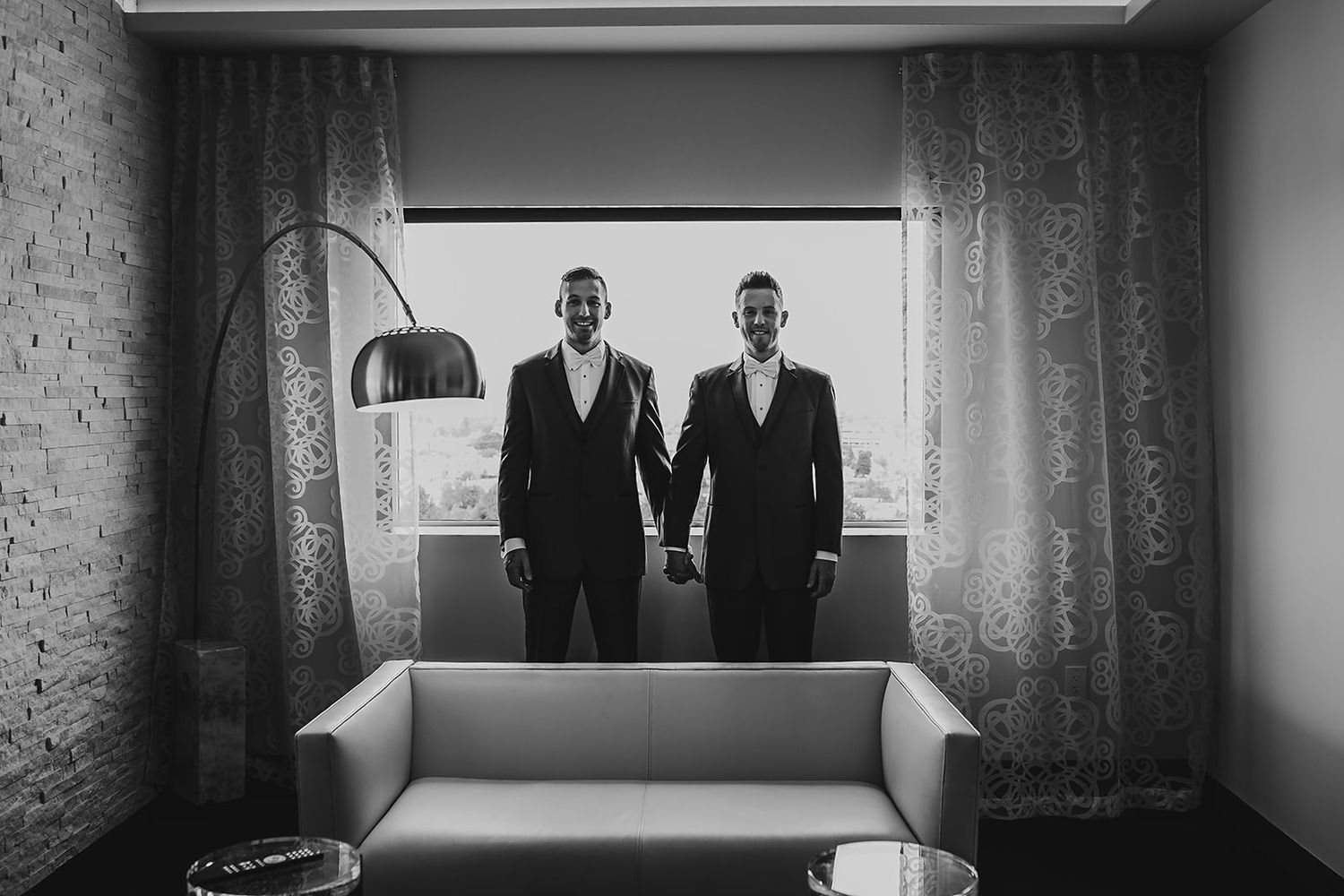 Two grooms in black suits hold hands smiling in a black and white wedding portrait.