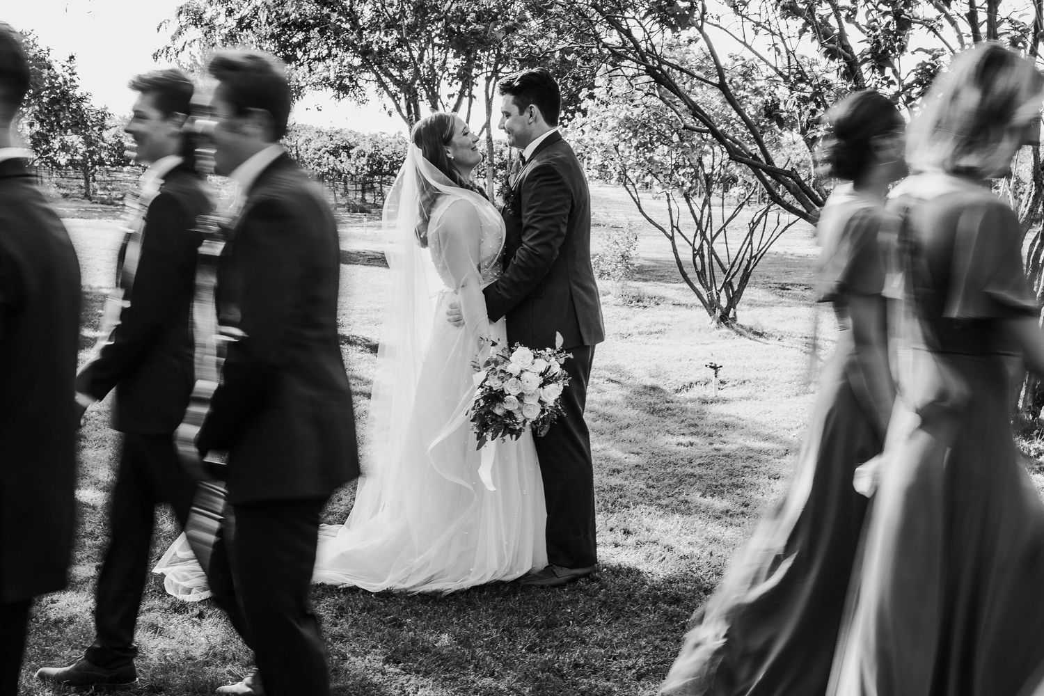 Black and white photo of bride and groom sharing a kiss while wedding party walks past in blur.
