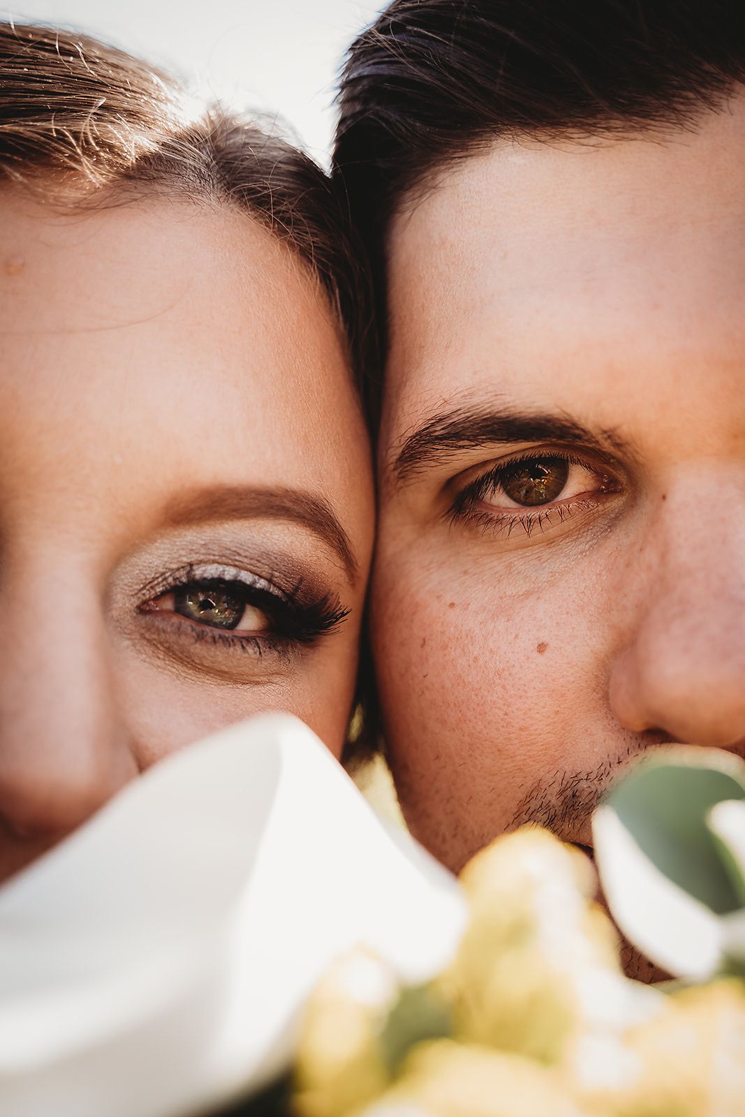 Close-up portrait of bride and groom faces together with green eyes visible above wedding bouquet.
