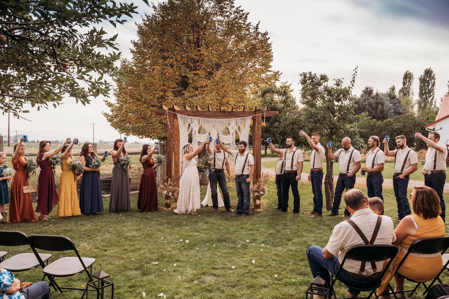Full wedding party raising drinks in celebration under wooden arch at outdoor boho ceremony.