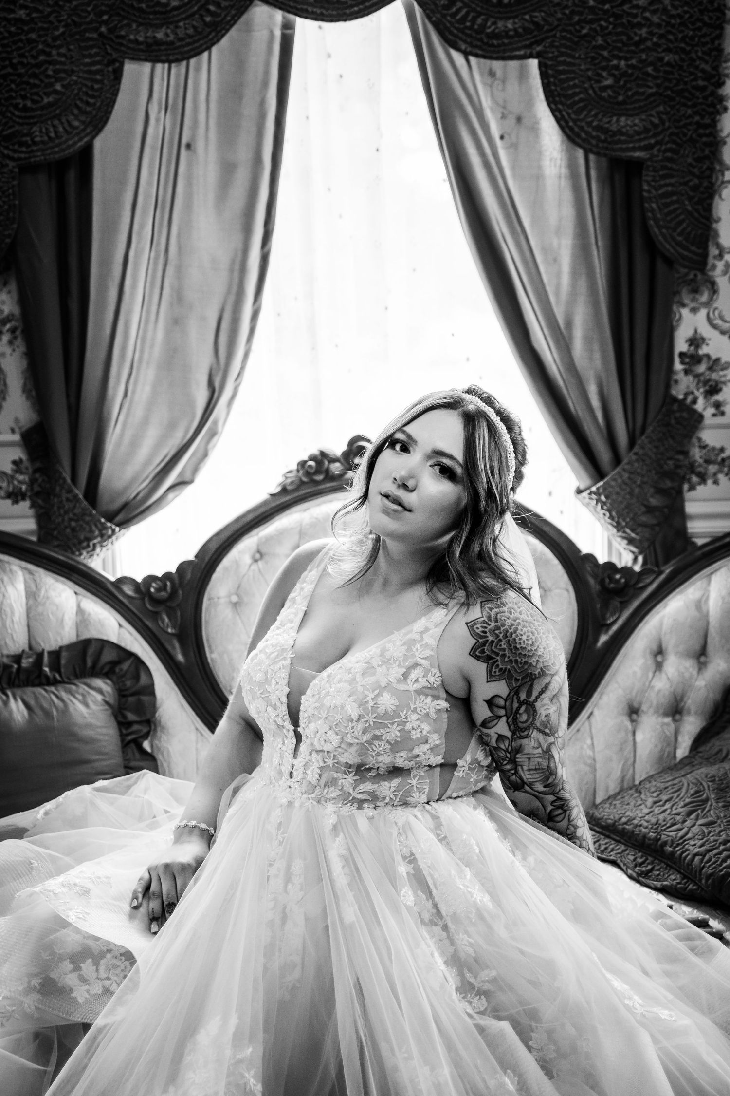Close-up of a tattooed bride in lace wedding dress seated on a vintage tufted sofa near a draped window.