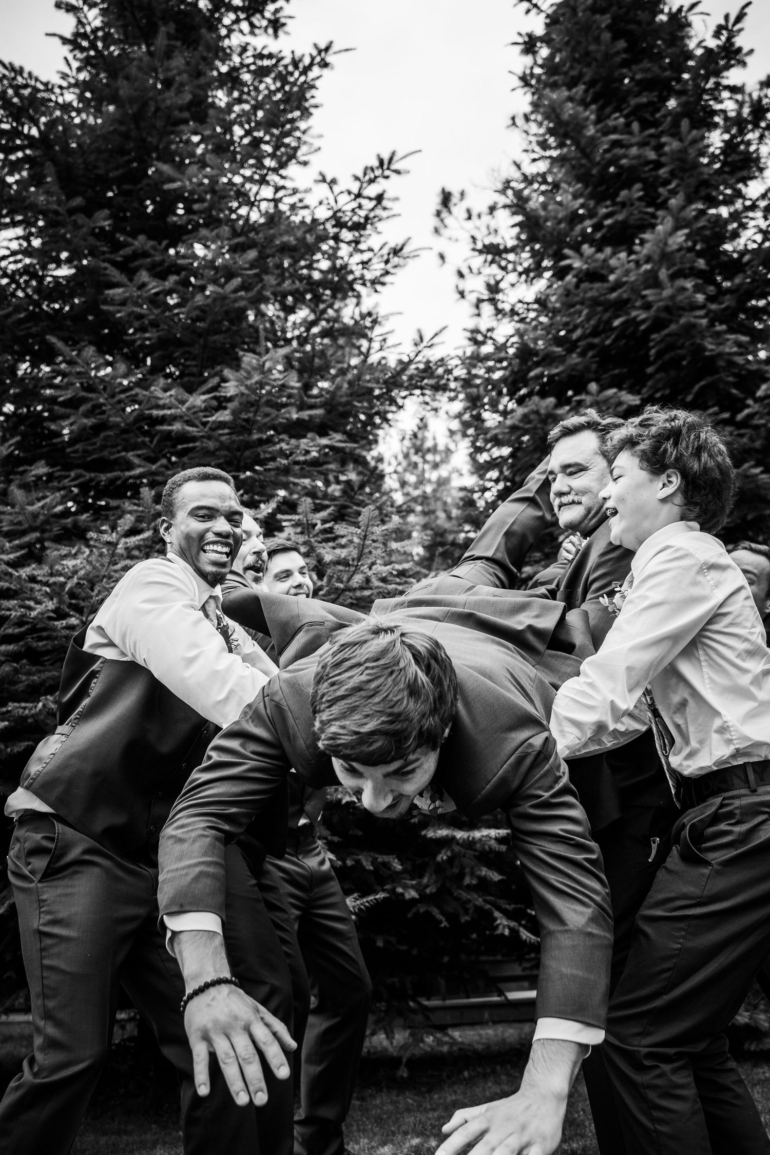 Groomsmen in suits playfully lifting the groom outdoors in a black and white wedding photo.