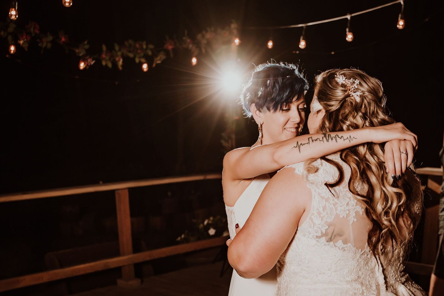 Two brides embrace during their first dance under glowing string lights at night.