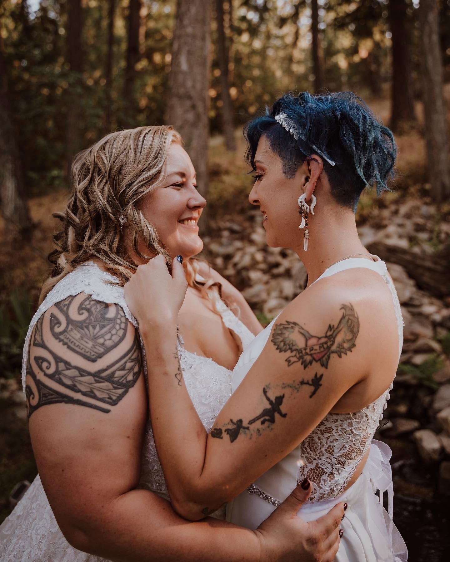 Two brides with tattoos embrace and smile at each other in a sunlit forest wedding setting.
