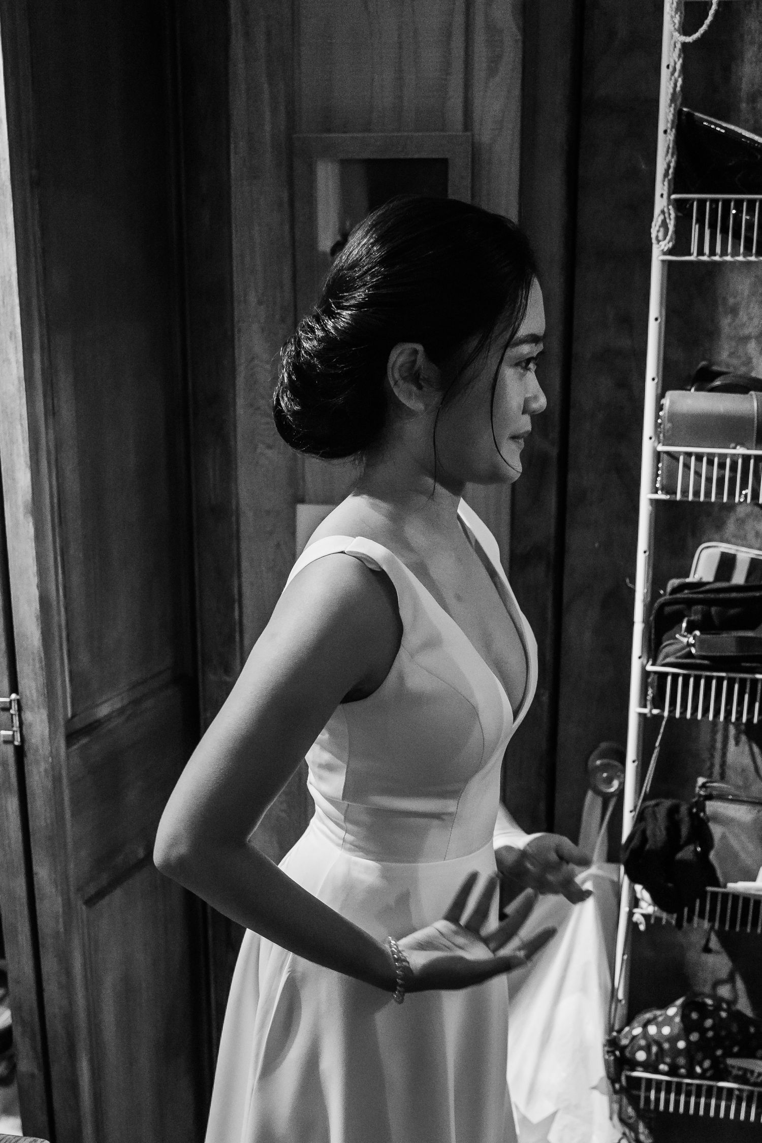 Elegant bride in white sleeveless gown with updo hairstyle standing near a closet in black and white photo.