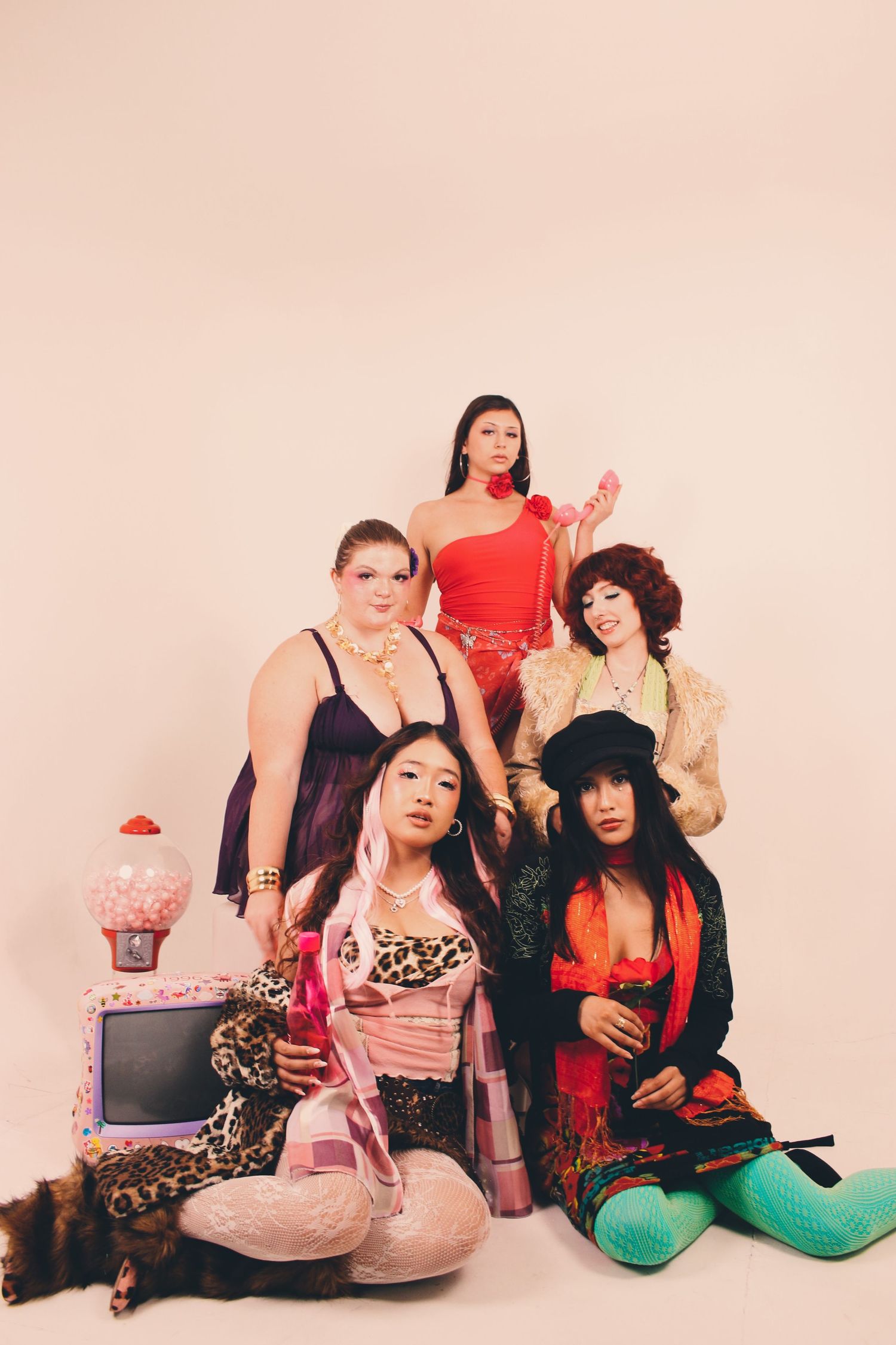 Five stylishly dressed women pose together in a studio, wearing colorful outfits with bold accessories and props.