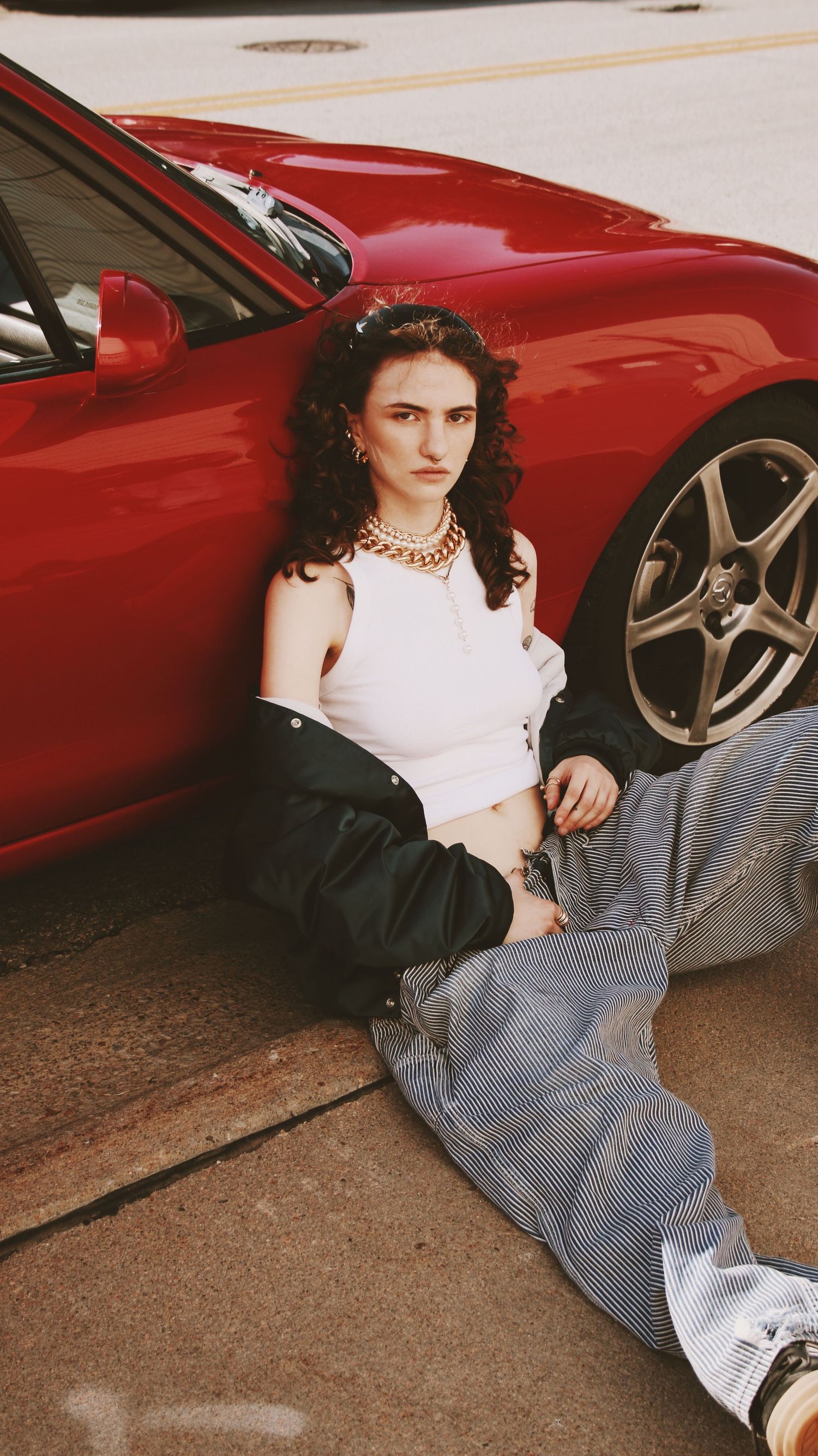 Young woman with curly hair sits against a red sports car, wearing a white crop top, black jacket, and wide-leg jeans.