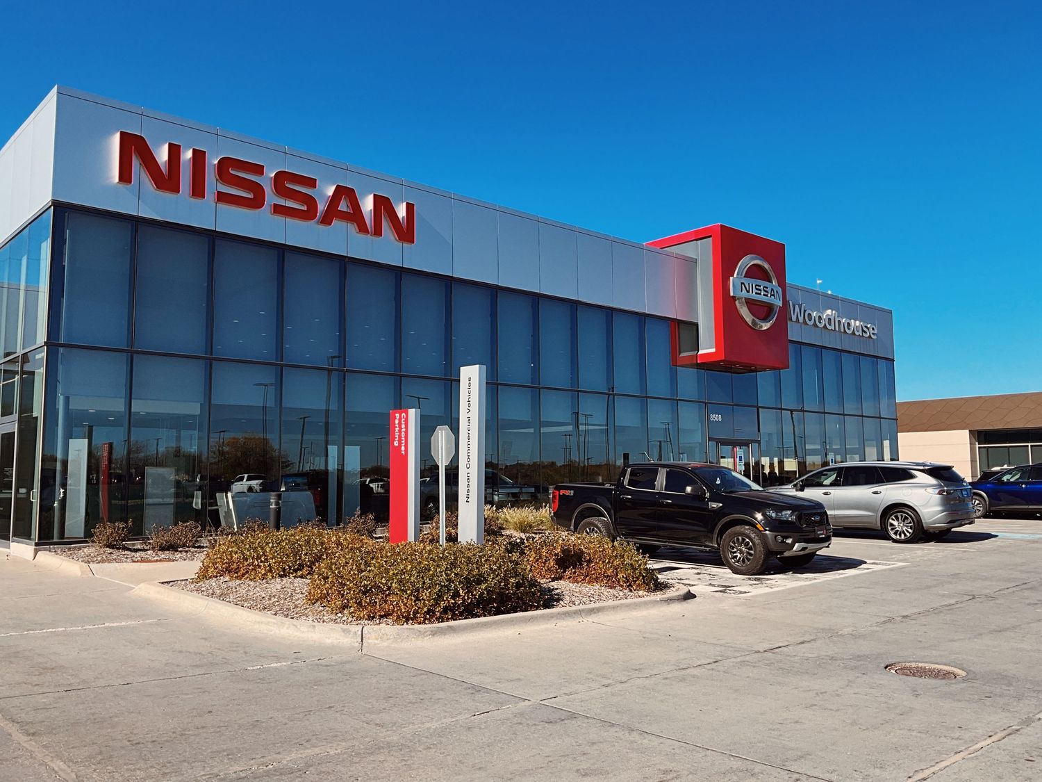 Nissan car dealership exterior with large red logo signage, glass facade, and vehicles parked in lot under clear blue sky.