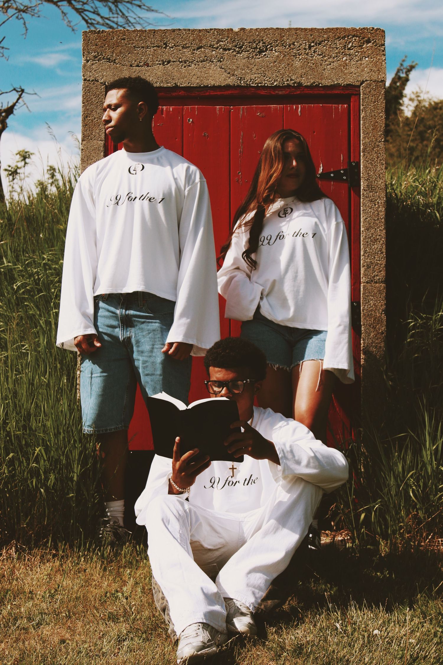 Three people in matching white oversized shirts pose by a red door outdoors, one reading a book.