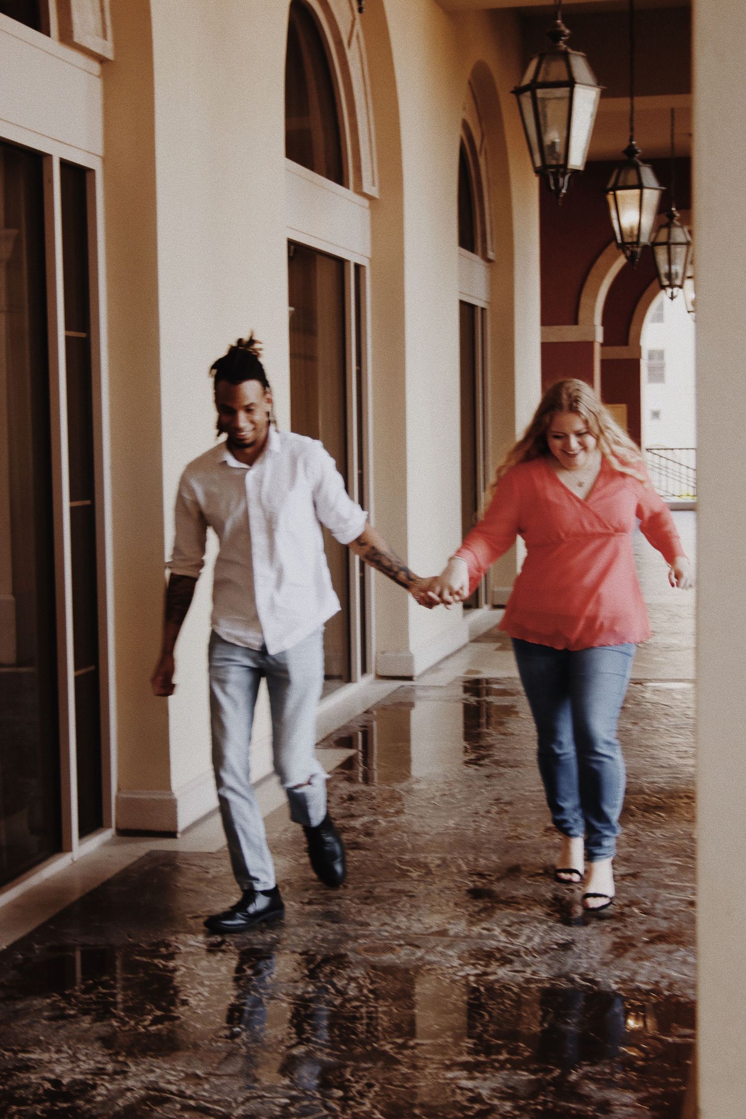Interracial couple holding hands and walking together under an elegant covered walkway with arched columns and lanterns.
