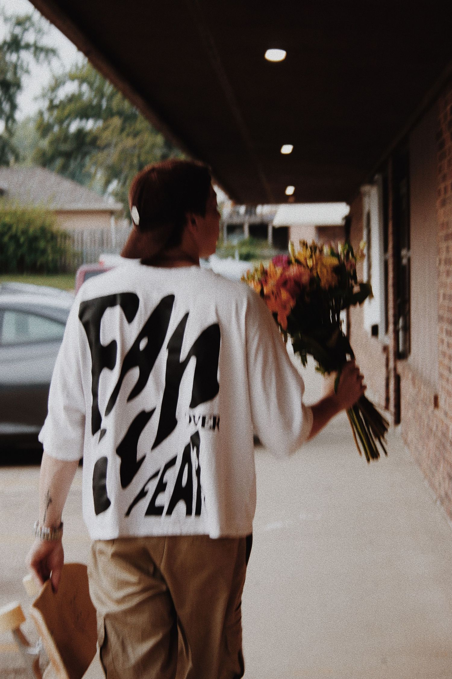 Person walking outdoors holding colorful flower bouquet, wearing white graphic tee with bold text and khaki pants.