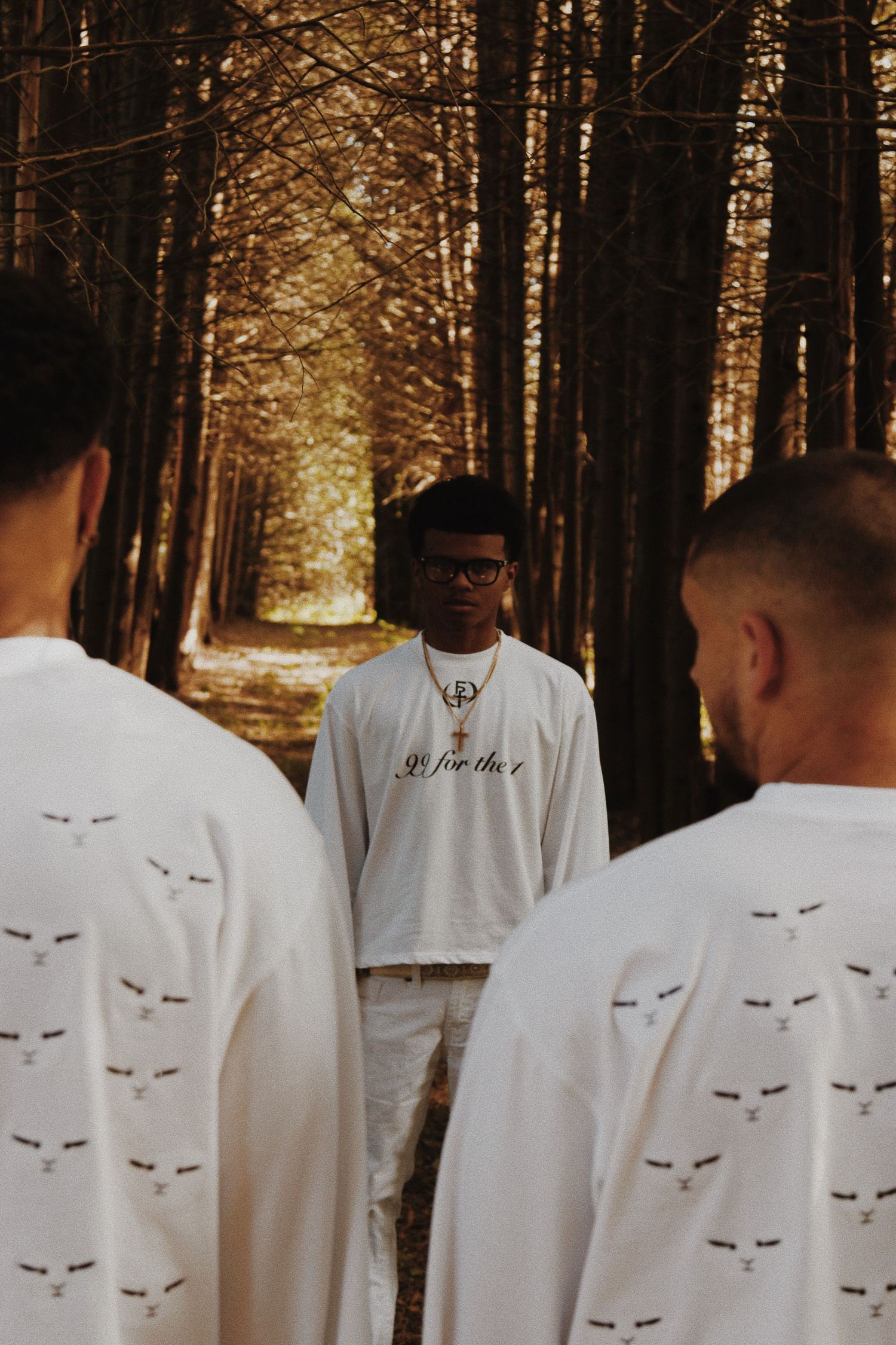 Young man in white graphic tee stands between two others in a golden sunlit forest path.