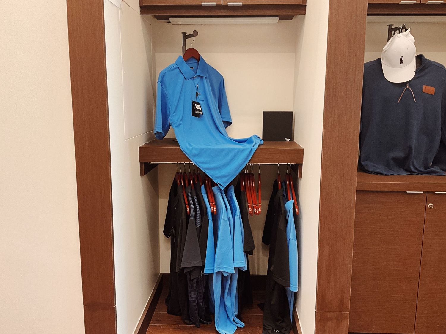 A retail store closet display with blue polo shirts and dark clothing hanging on wooden racks with a white cap on shelf.