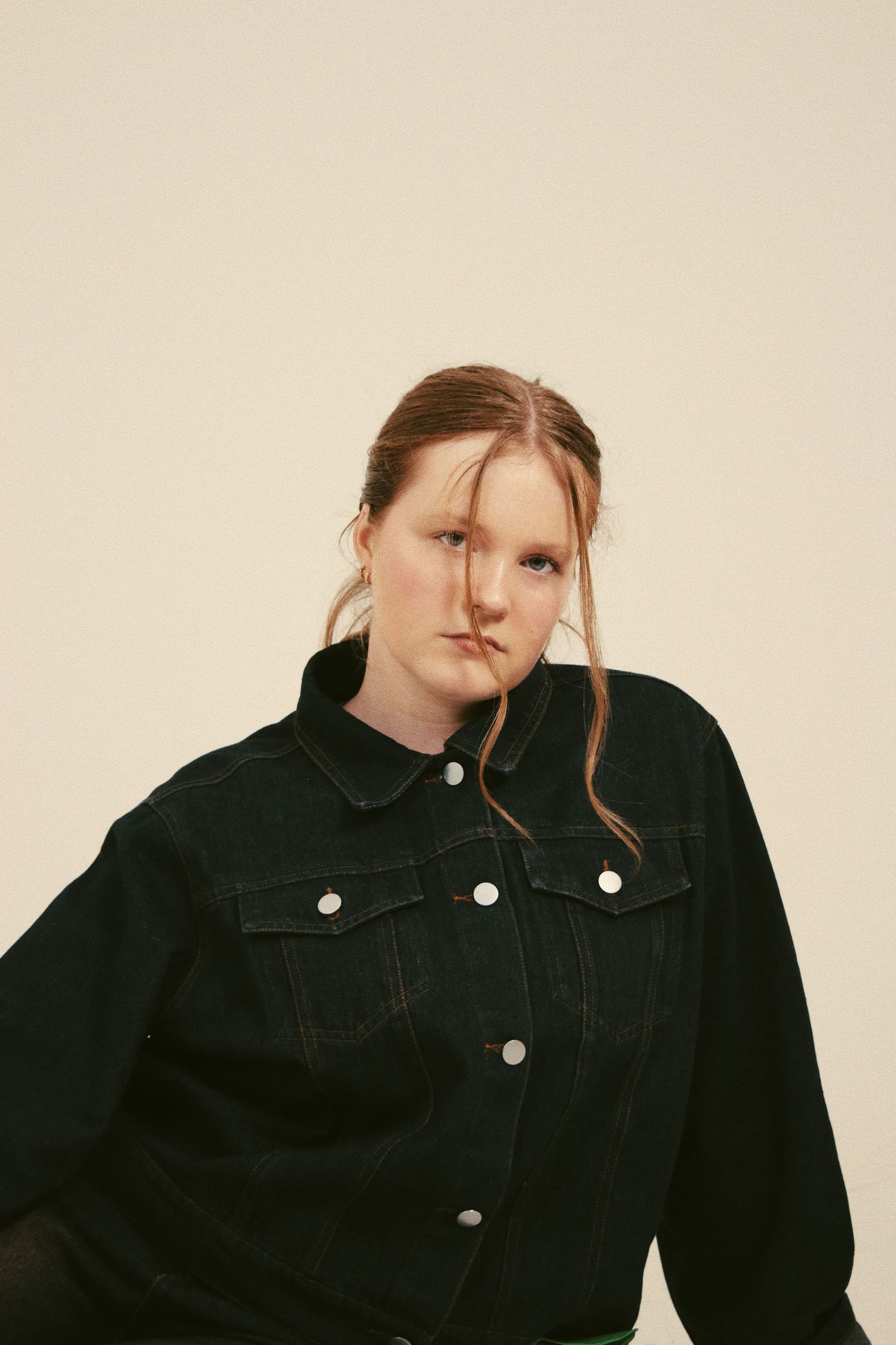 Young woman in black denim jacket with hair across face poses against light background.