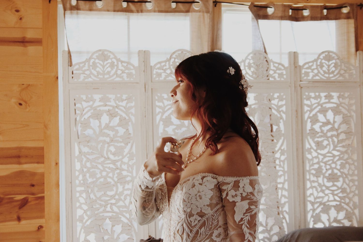 Bride in lace off-shoulder gown holding perfume bottle by a decorative white curtained window in a wooden room.