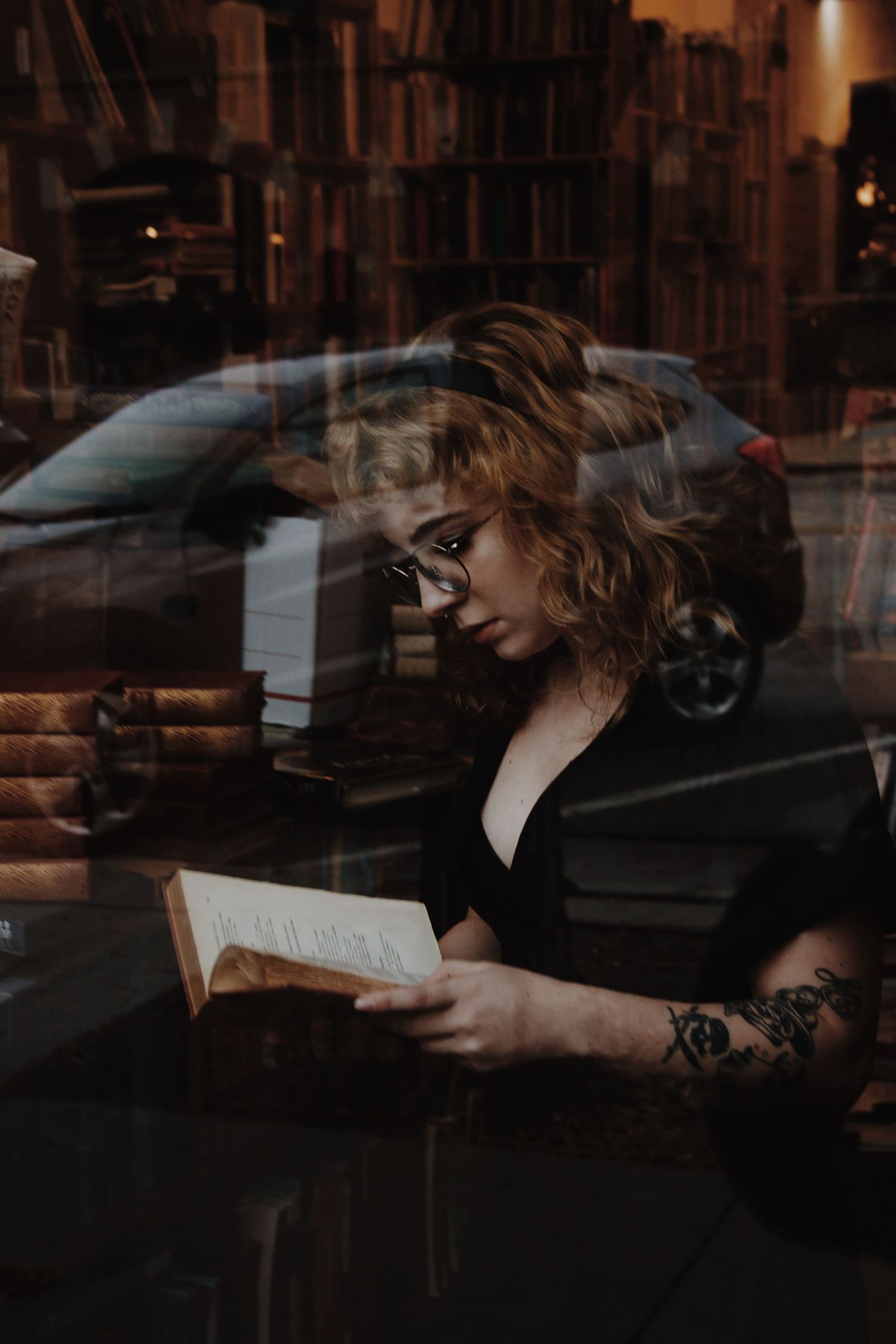 Young woman with curly hair and tattoos reading a book in a cozy bookstore, viewed through a window.