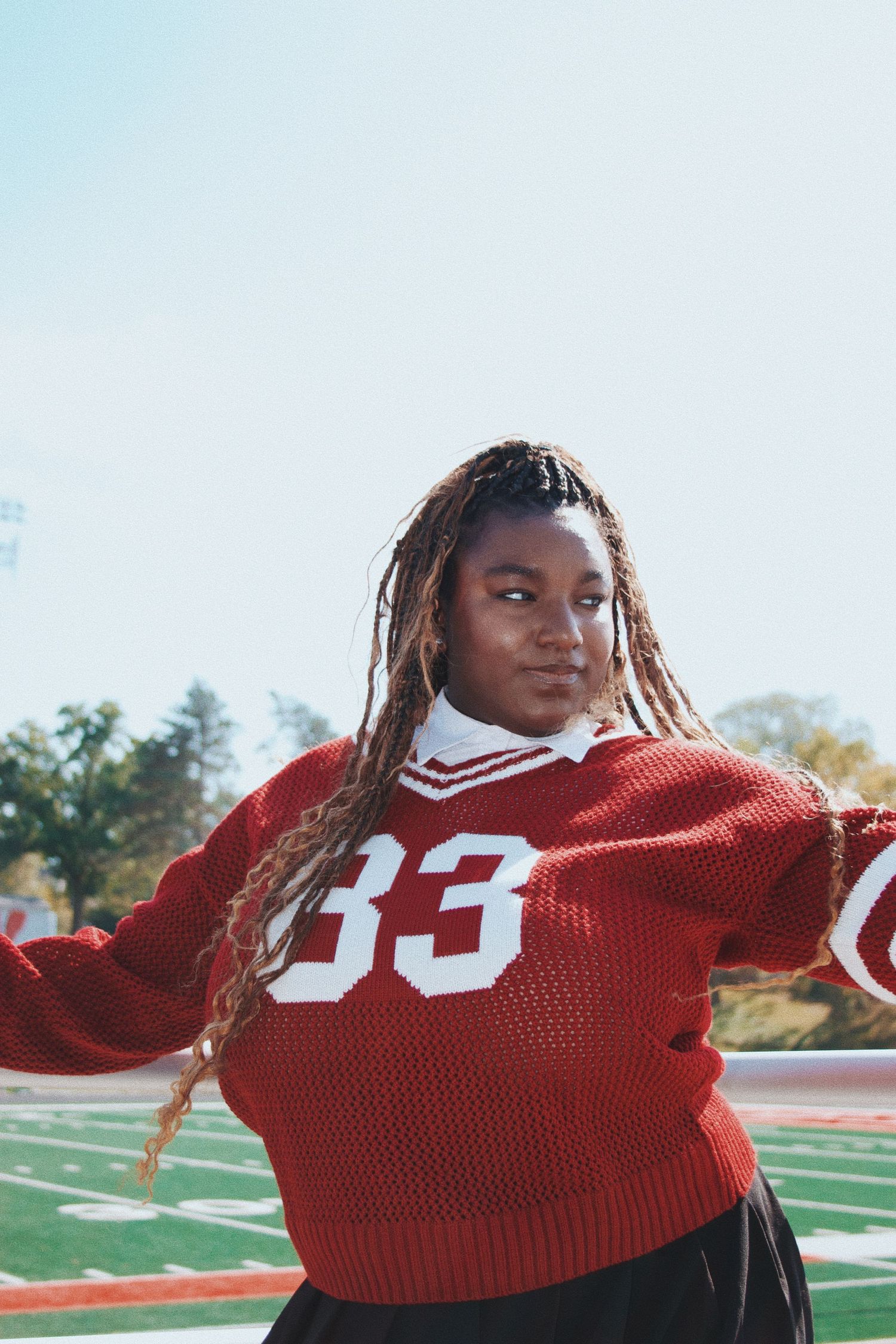 Young woman wearing red number 33 varsity sweater posing on green football field on sunny day.