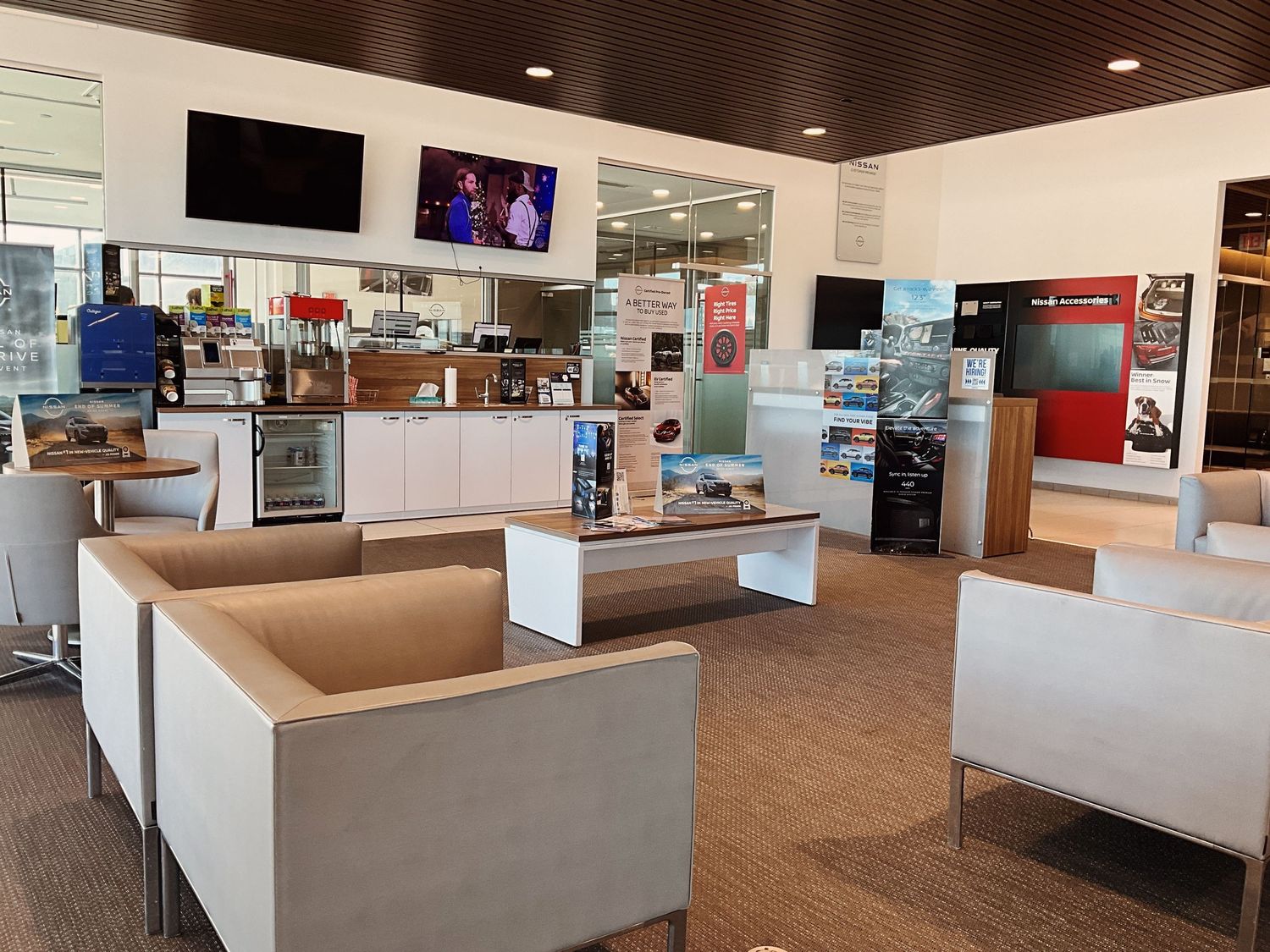 Modern car dealership lounge with white furniture, flat-screen TVs, and a refreshment bar area.