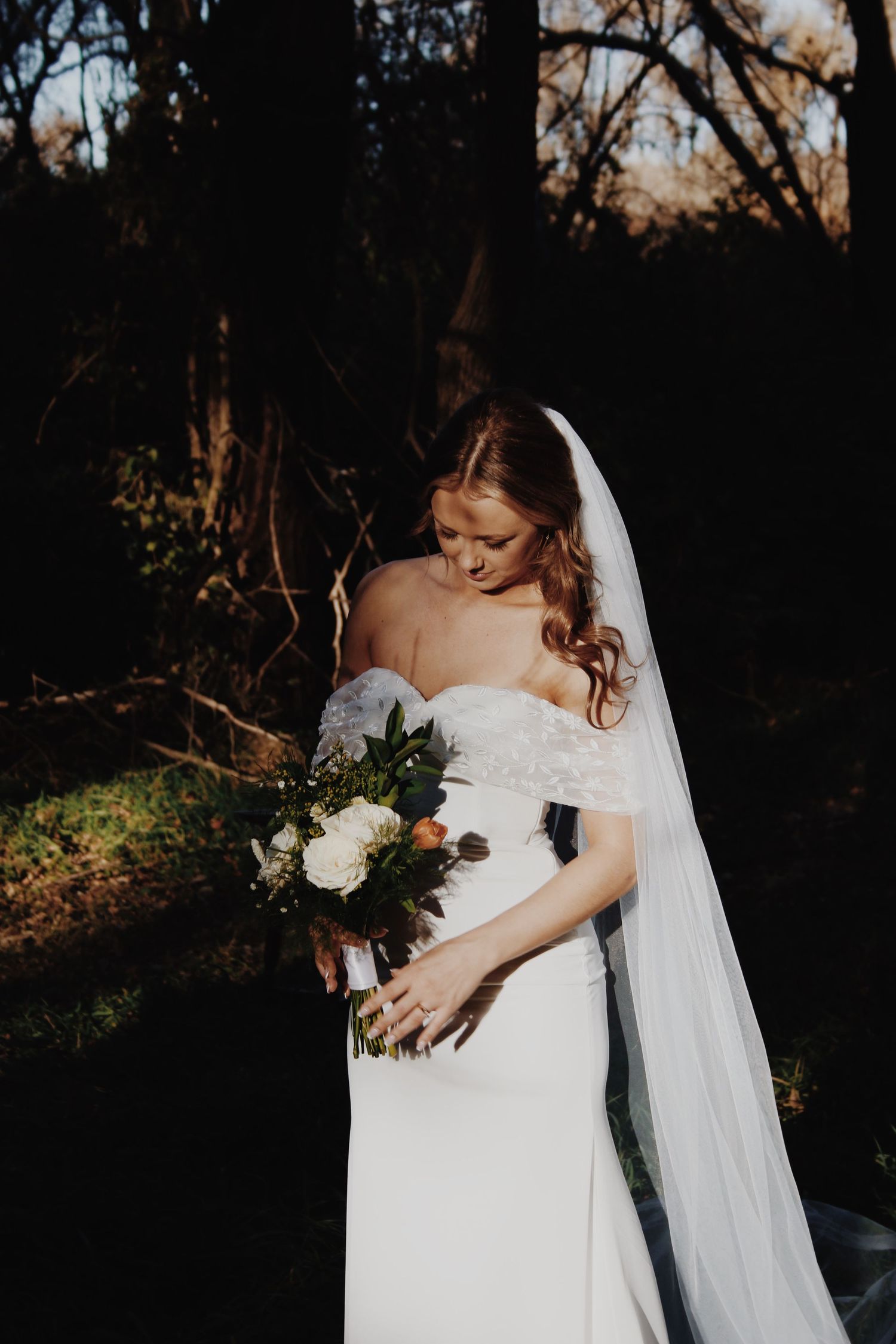 Bride in off-shoulder white gown and veil holds floral bouquet outdoors in dappled sunlight.