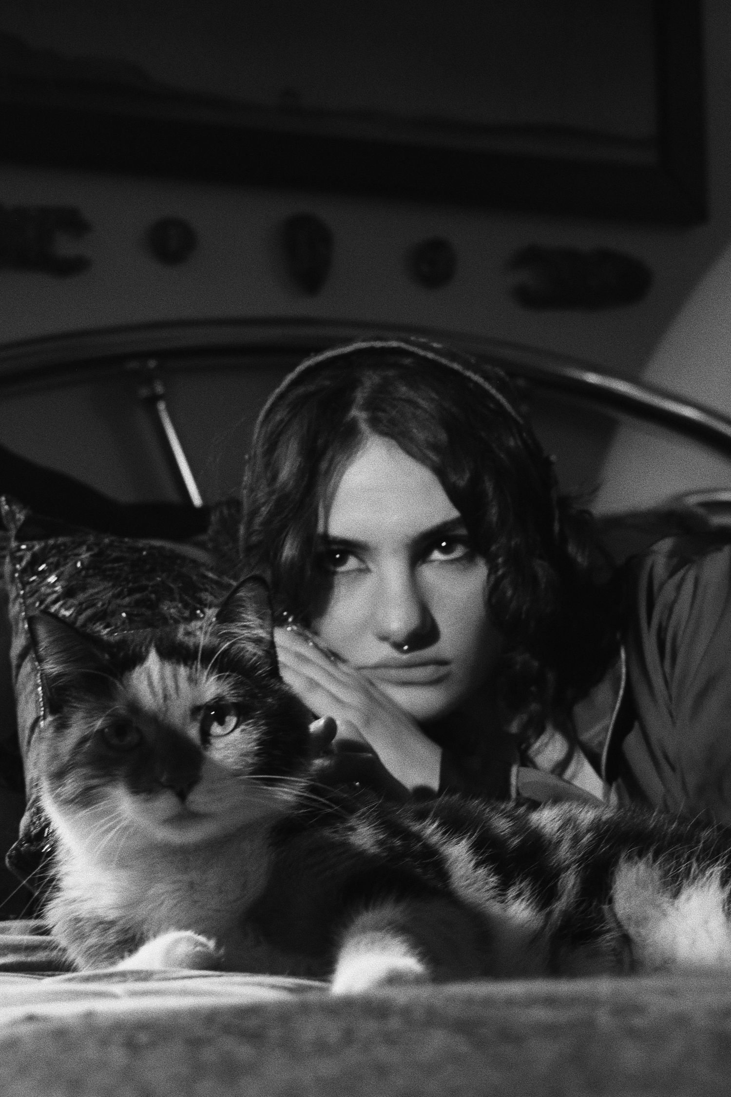 Black and white portrait of a young woman resting her cheek on her hand beside a fluffy calico cat on a couch.