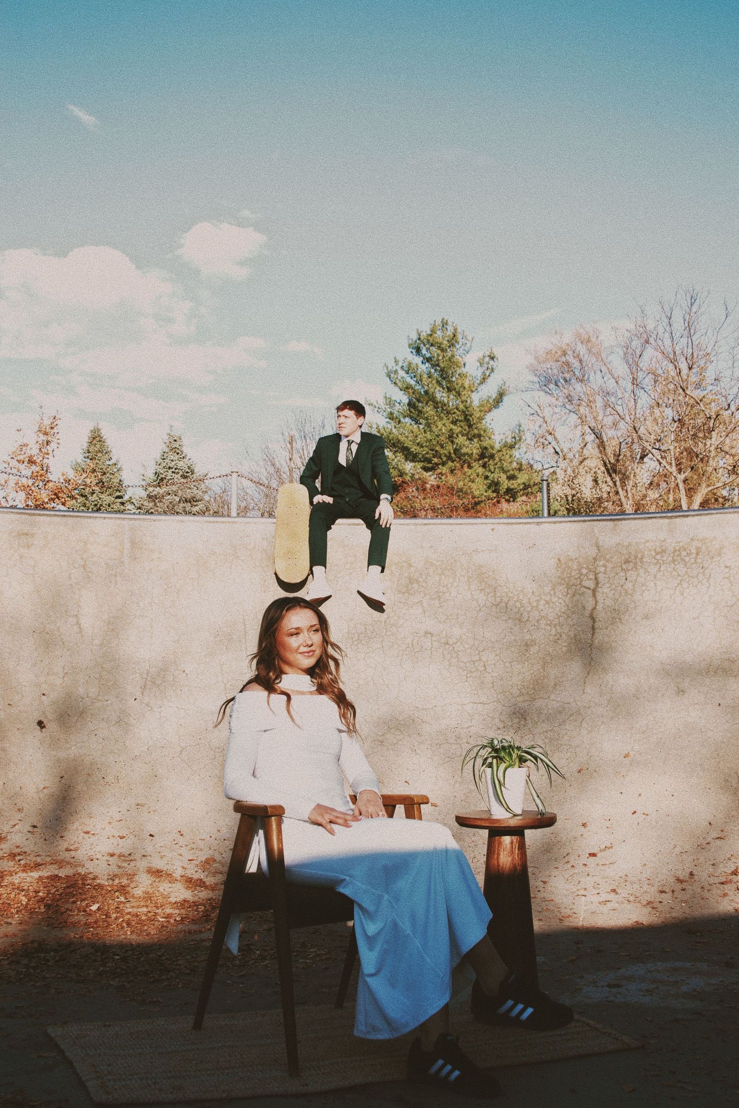 A bride in a white top and blue skirt sits in a wooden chair while a suited groom perches on a wall behind her outdoors.