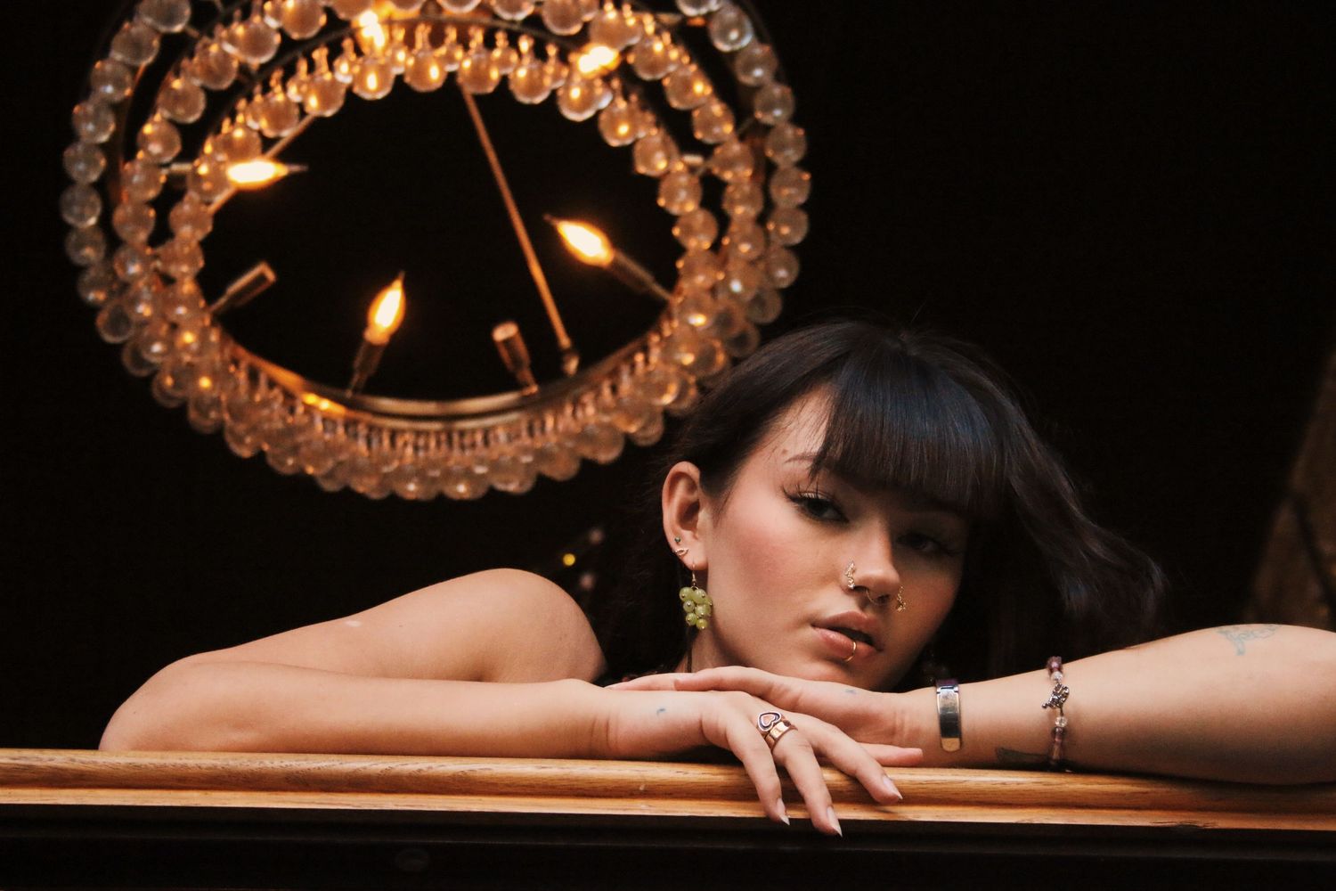 A woman with dark hair rests her chin on a wooden surface beneath a glowing crystal chandelier in a dark setting.
