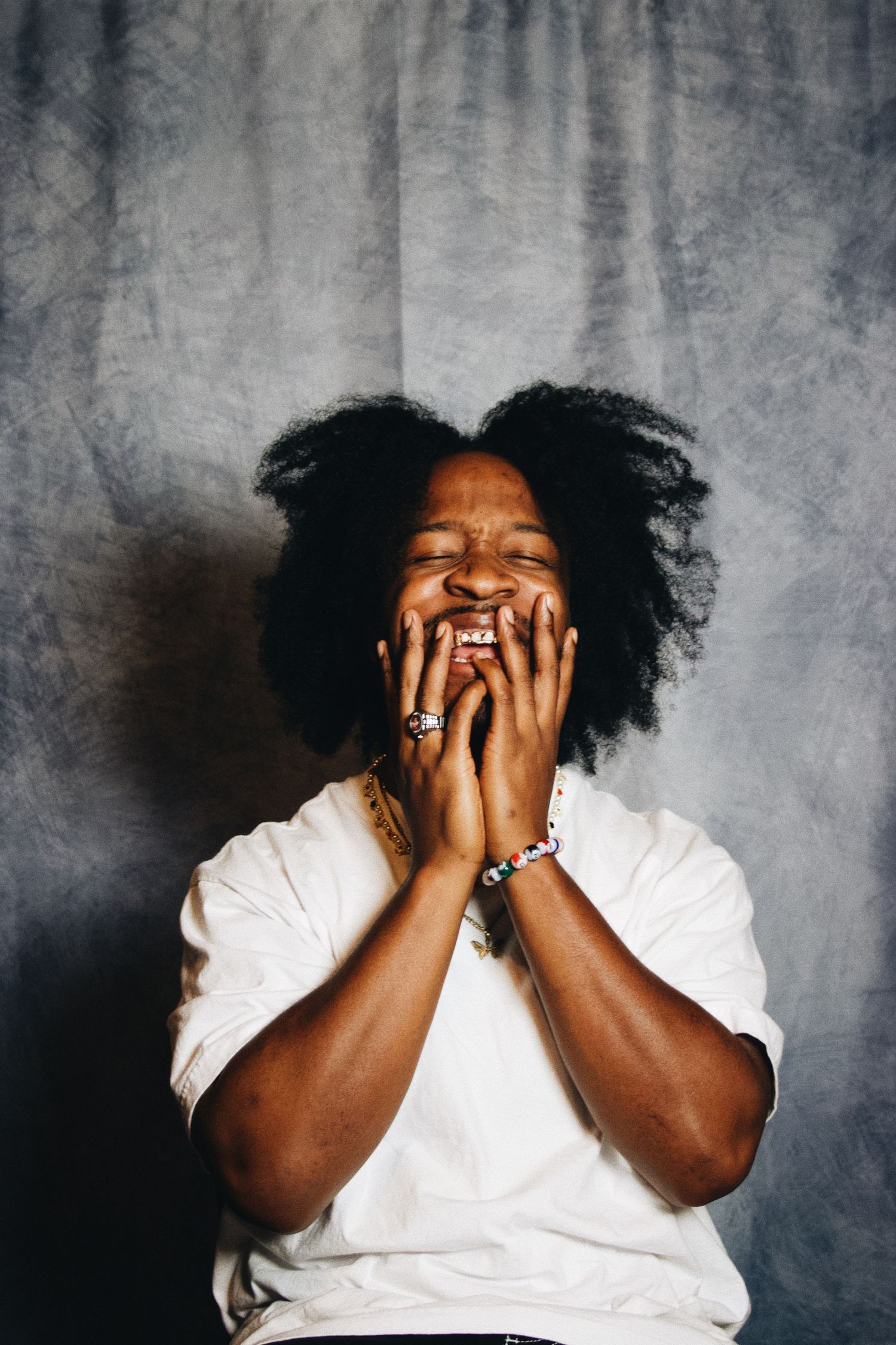 A joyful person with natural afro hair laughing with hands on face, wearing a white tee and bracelet against gray backdrop.