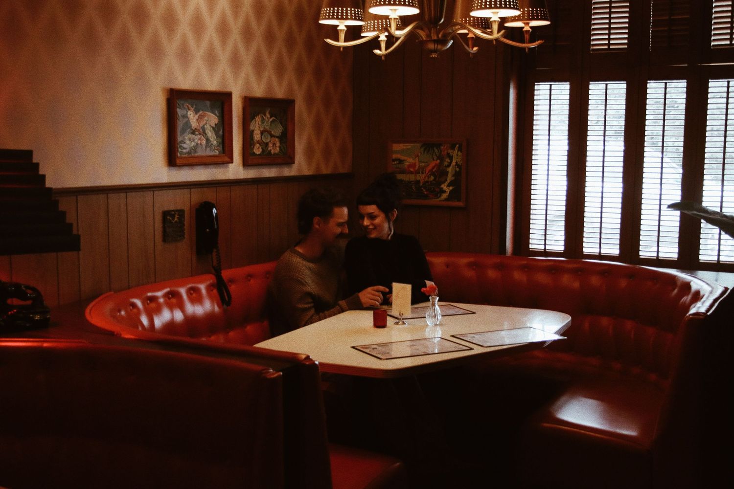 Couple sharing an intimate moment in a cozy red leather booth at a dimly lit vintage restaurant.