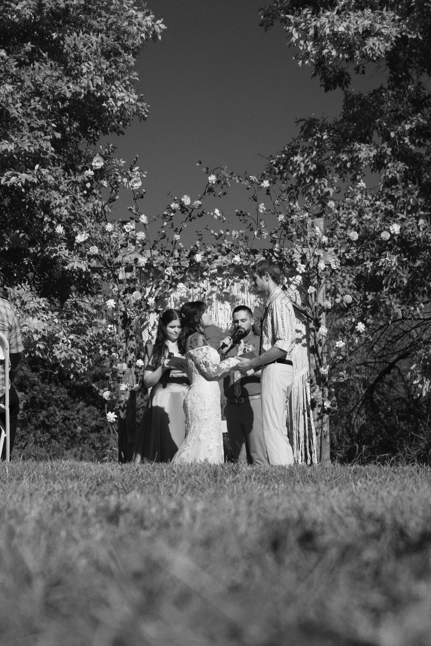 Black and white outdoor wedding ceremony with couple exchanging vows under floral arch surrounded by trees.