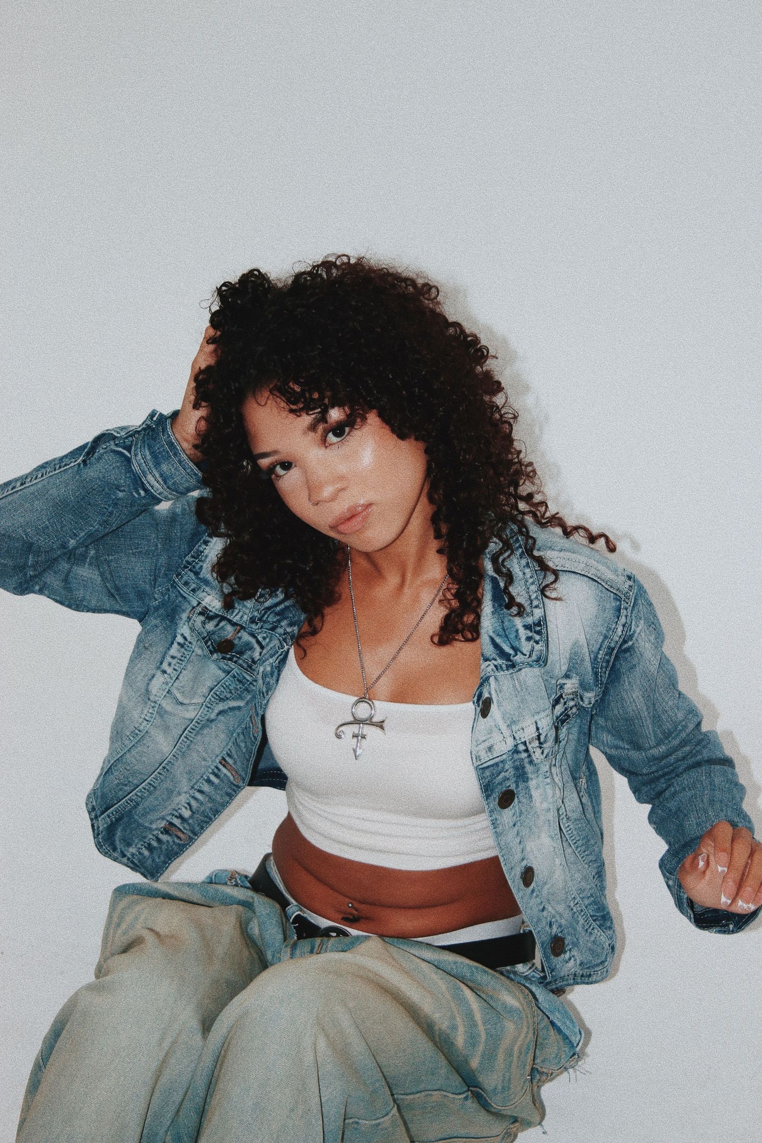 Young woman with curly hair in denim jacket, white crop top, and wide-leg jeans posing against white background.