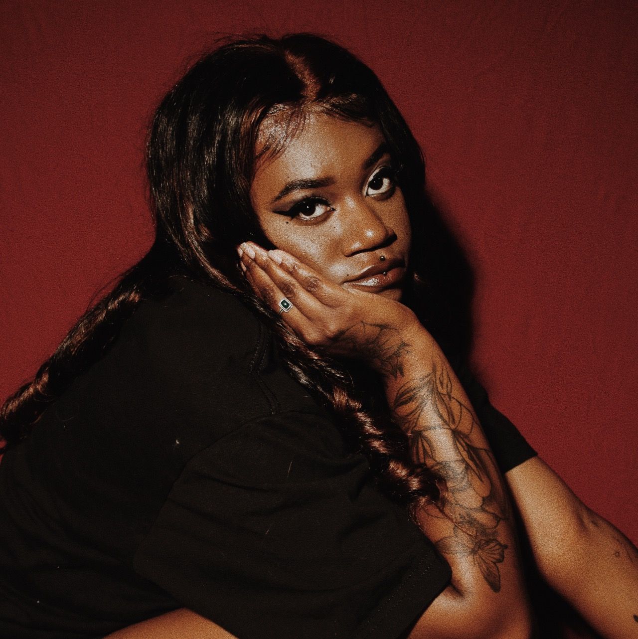 Young woman with tattoos on her arm poses confidently against a deep red background in a black outfit.