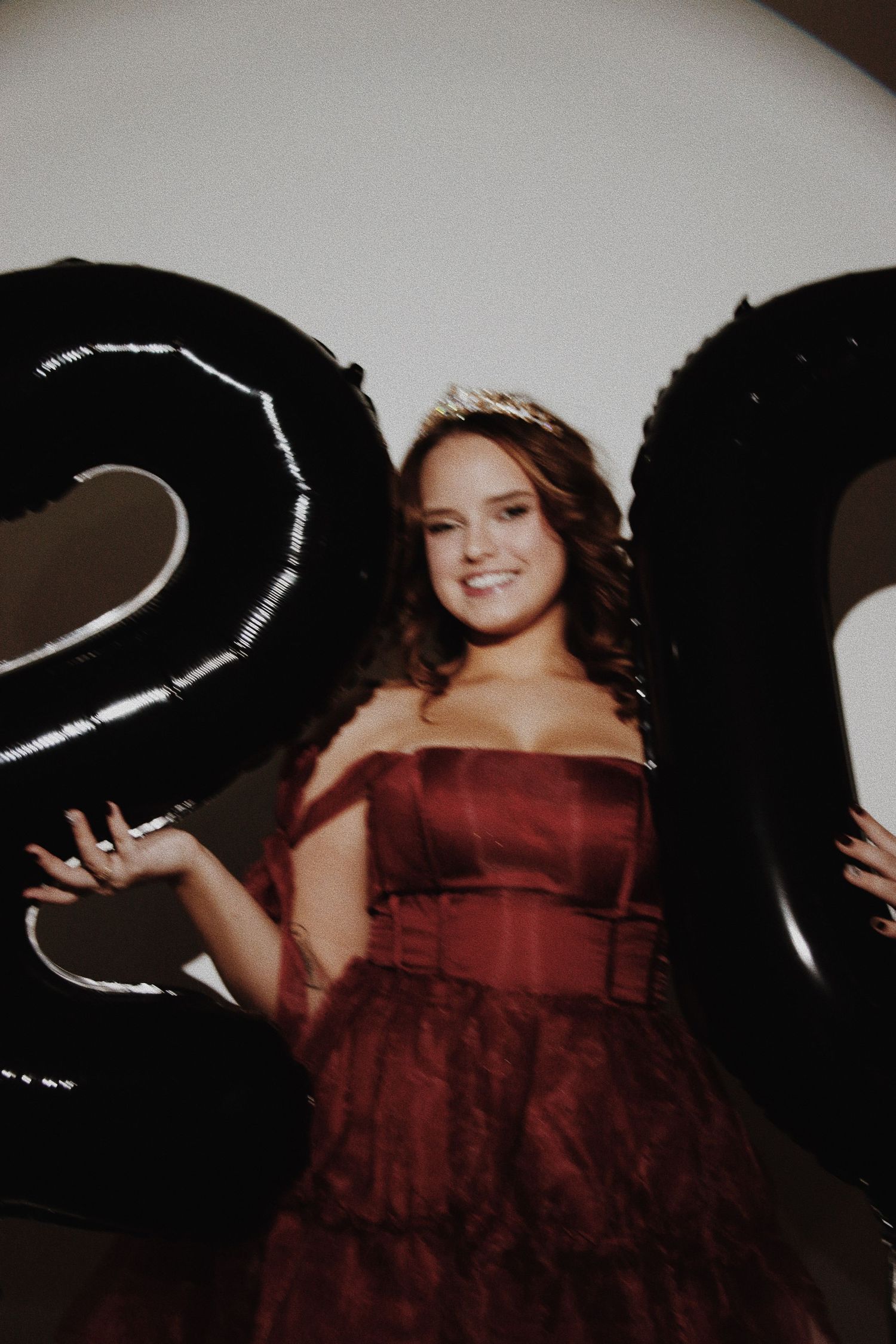 Young woman in burgundy off-shoulder gown wearing tiara, posing between black number balloons at birthday celebration.