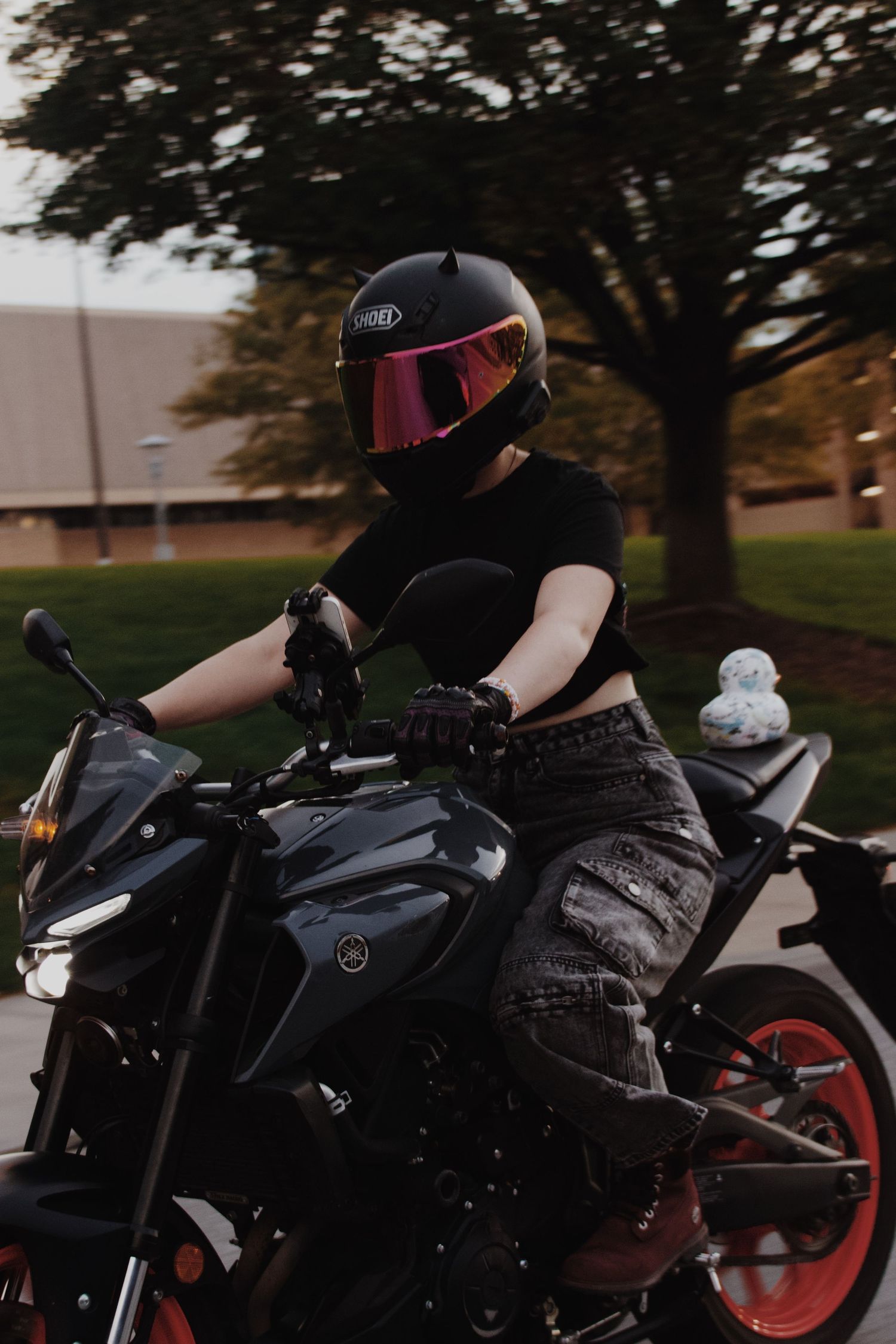 A woman in a black helmet with pink visor rides a dark Yamaha motorcycle outdoors near trees at dusk.