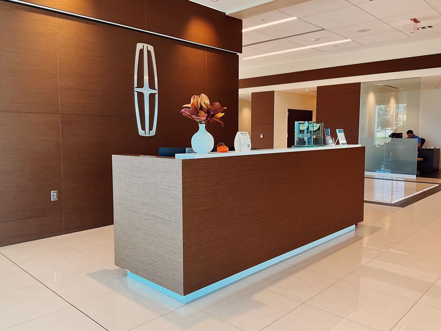 Modern car dealership reception desk with brown wood paneling, blue LED lighting, and Lincoln logo on wall.