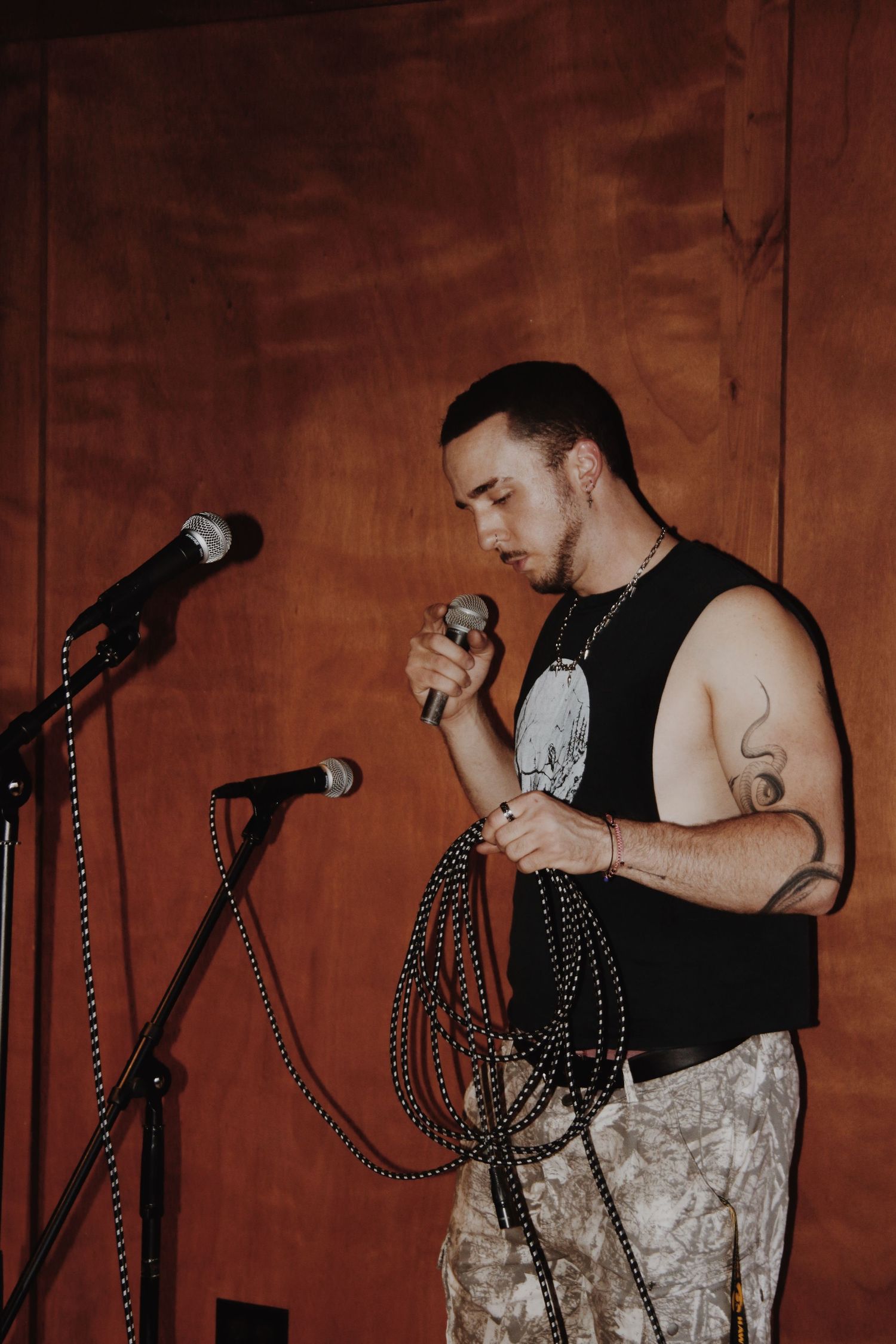 A tattooed male singer holds a microphone and cables while performing on stage near a mic stand.