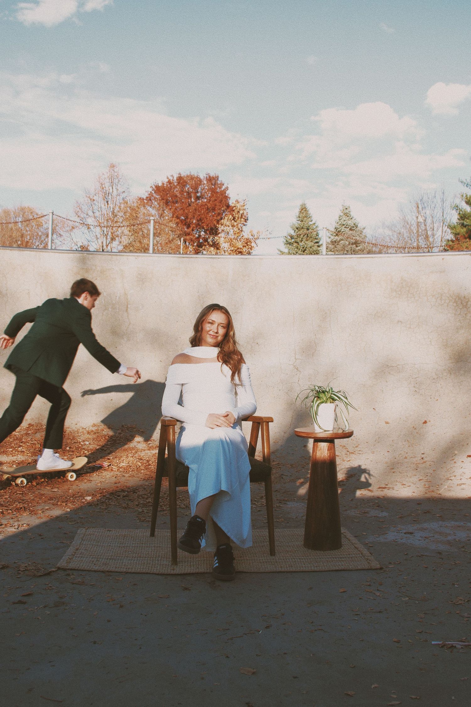 Pregnant woman in white sweater and blue skirt sits outdoors while man skateboard behind her near autumn trees.