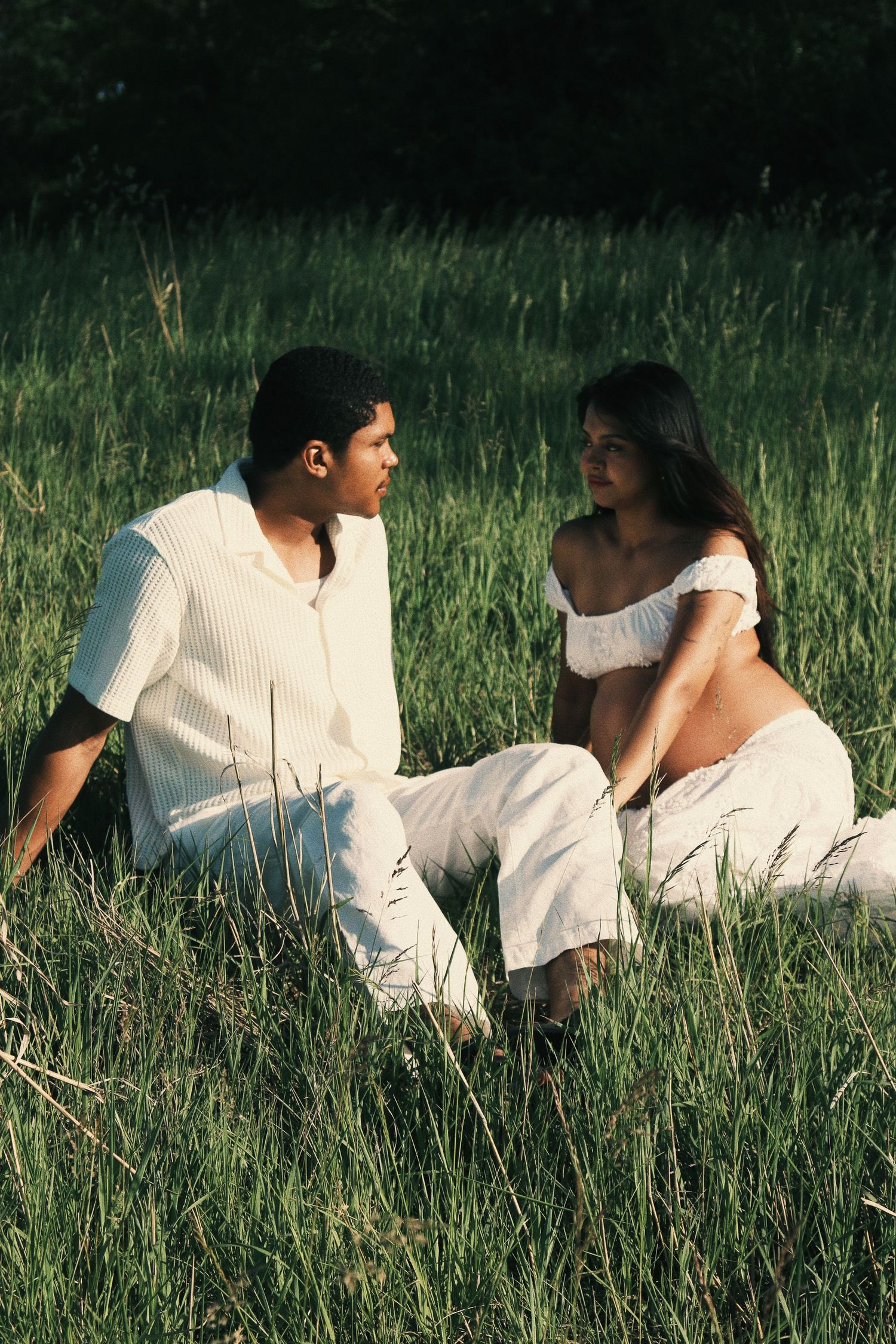 Young couple in white outfits sitting together in a lush green field, sharing an intimate conversation.