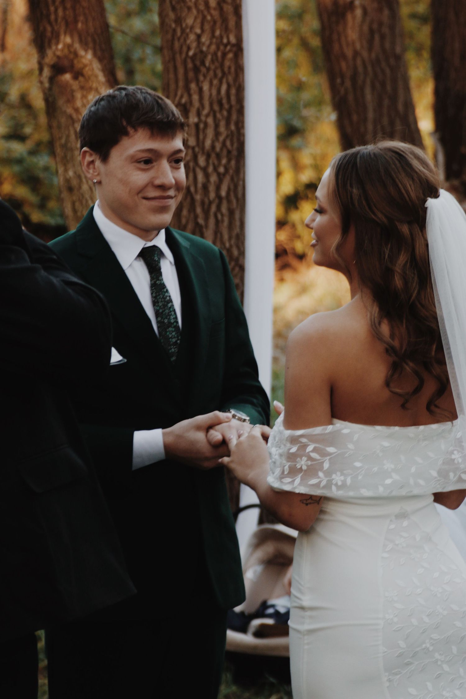 Groom in green velvet suit exchanges rings with bride in lace off-shoulder gown at outdoor ceremony.